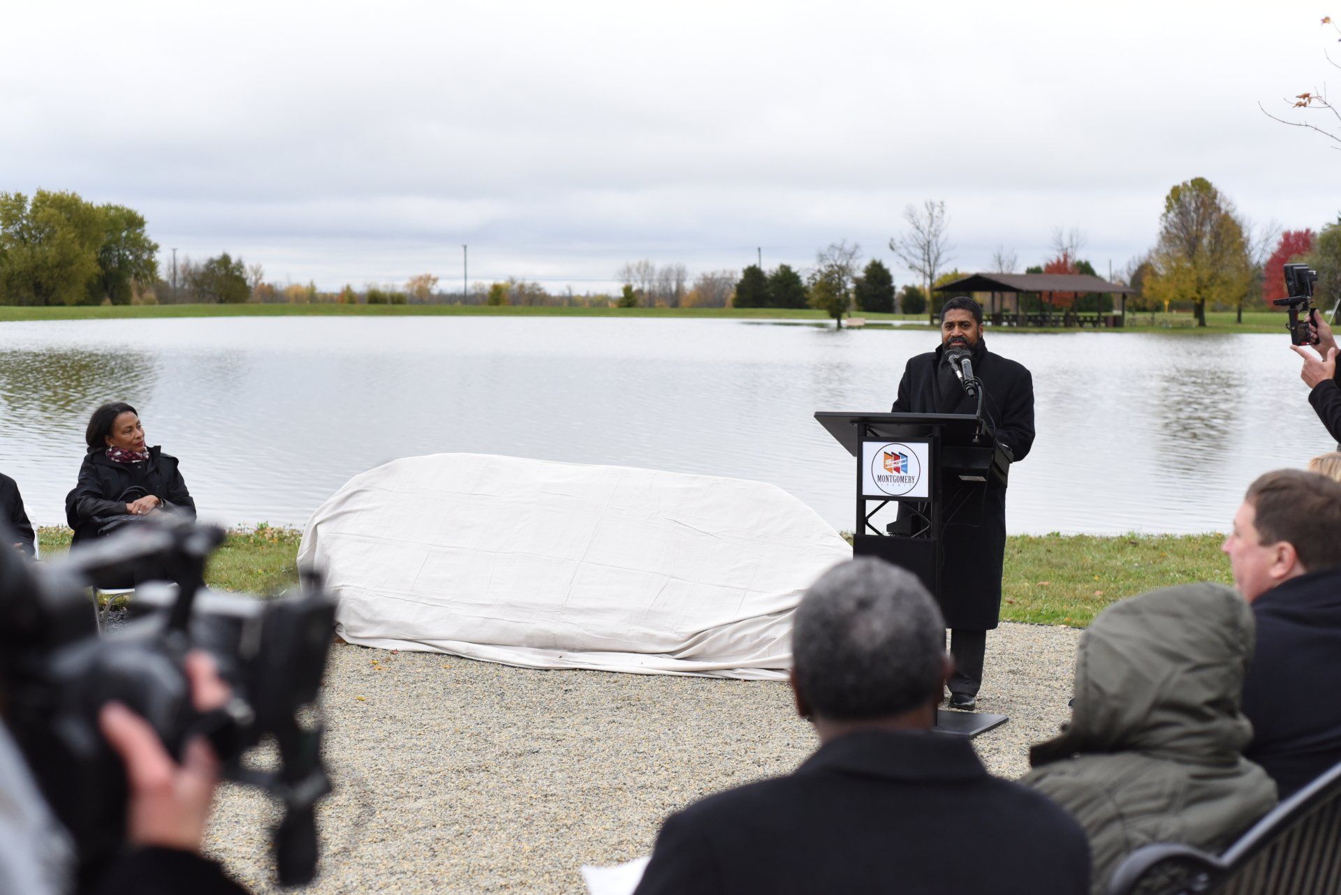 Man speaking at a podium near a pond, unveiling a covered stone, with an audience watching. Overcast day.