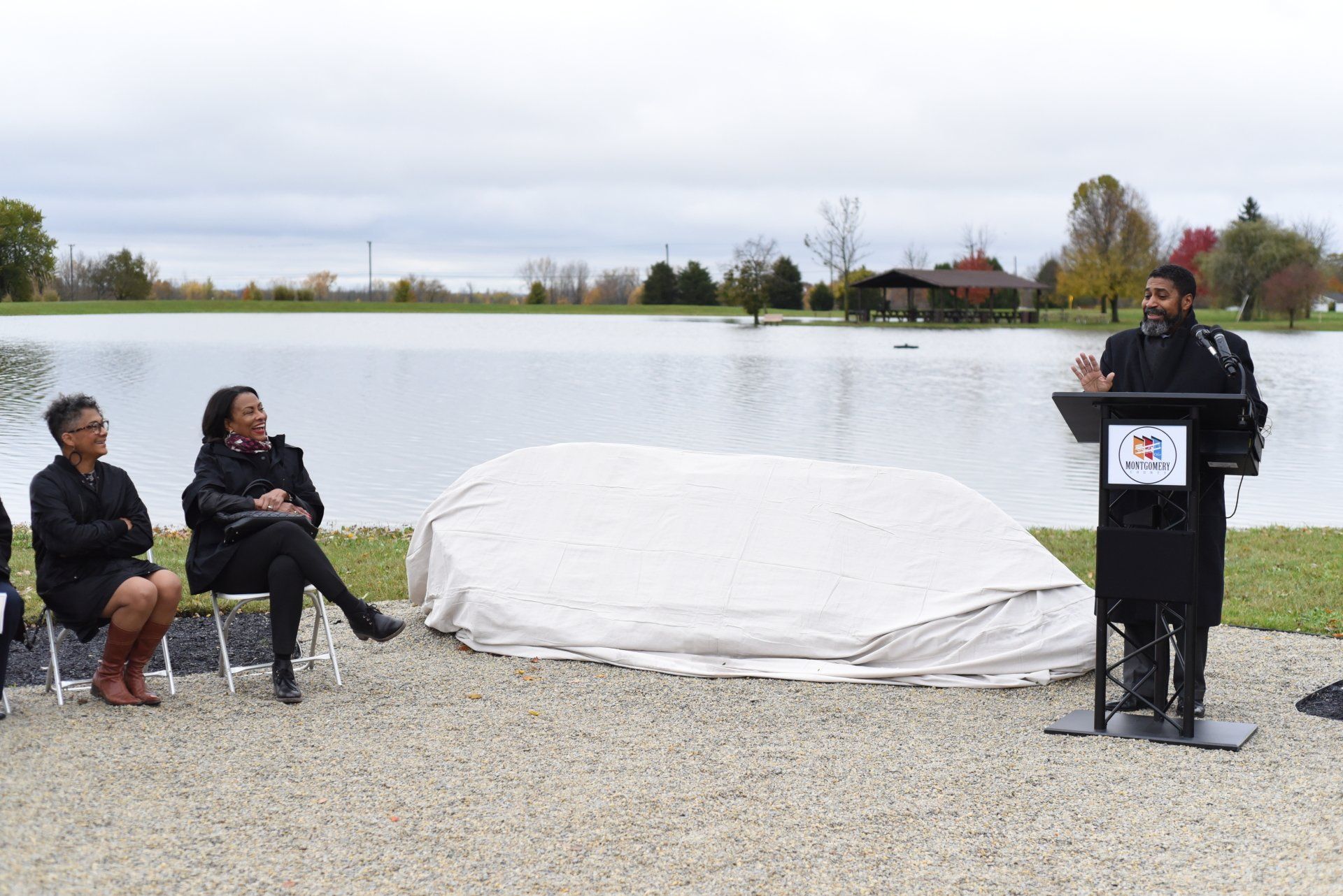 Man speaking at podium near a covered object, two women seated, a lake in the background on an overcast day.