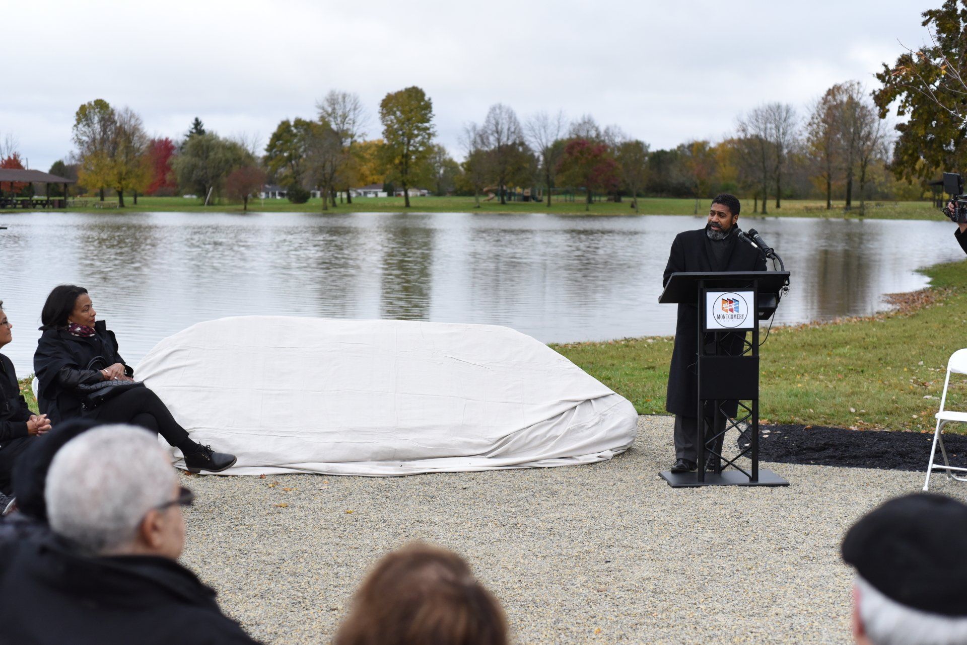 Man speaking at podium, covered object, people seated, lake backdrop on overcast day.