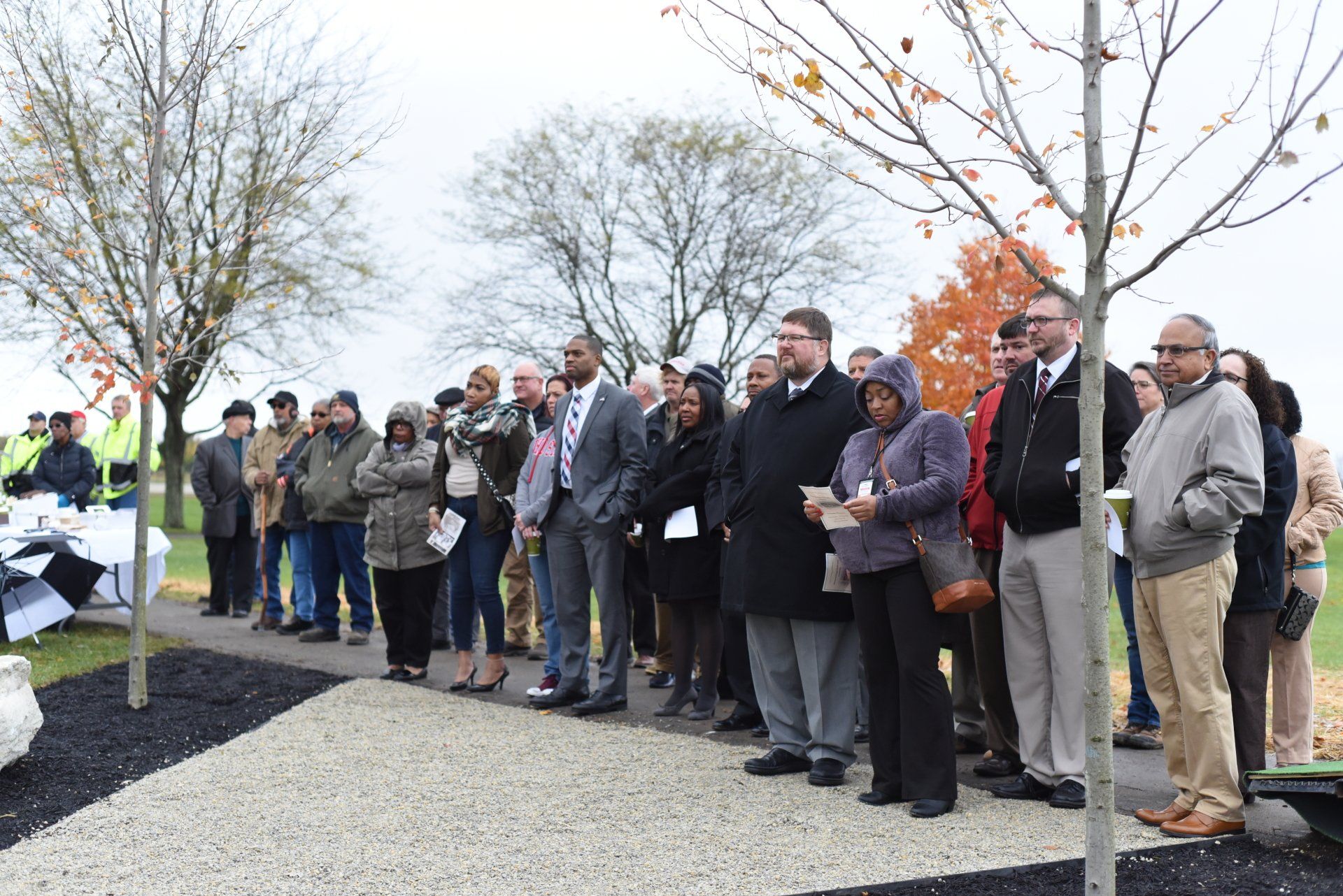 Group of people standing outdoors, listening to a speech, near trees with autumn leaves.