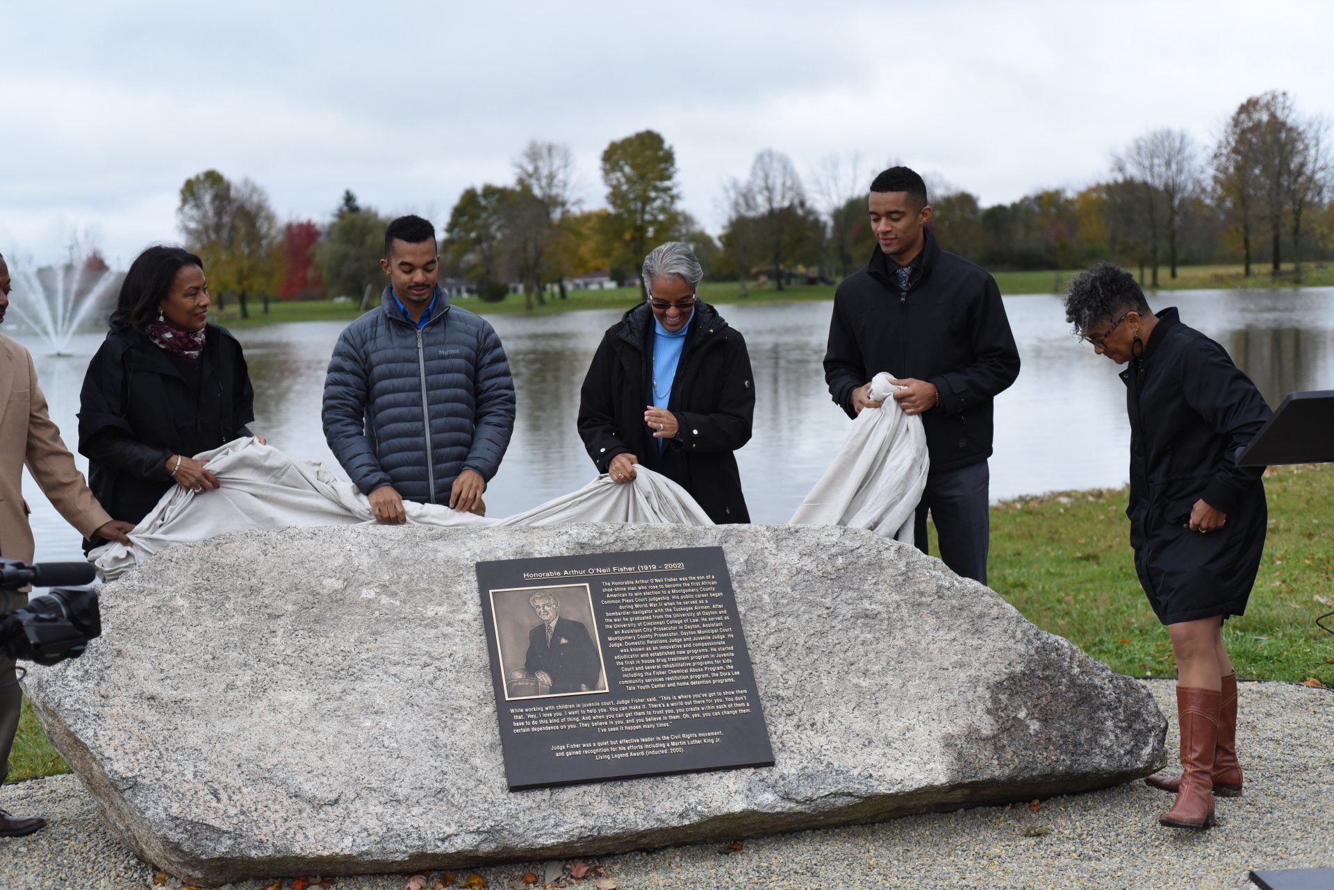 People unveil a memorial plaque on a stone by a lake. They are diverse and wearing coats. It’s a cloudy day.