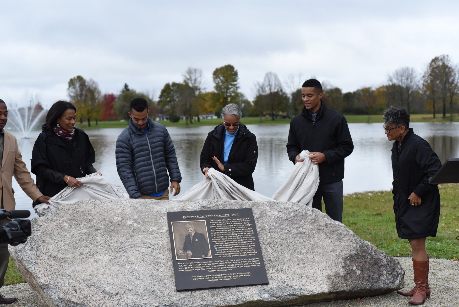 People unveiling a memorial plaque near a lake. Autumn setting with trees and fountain.