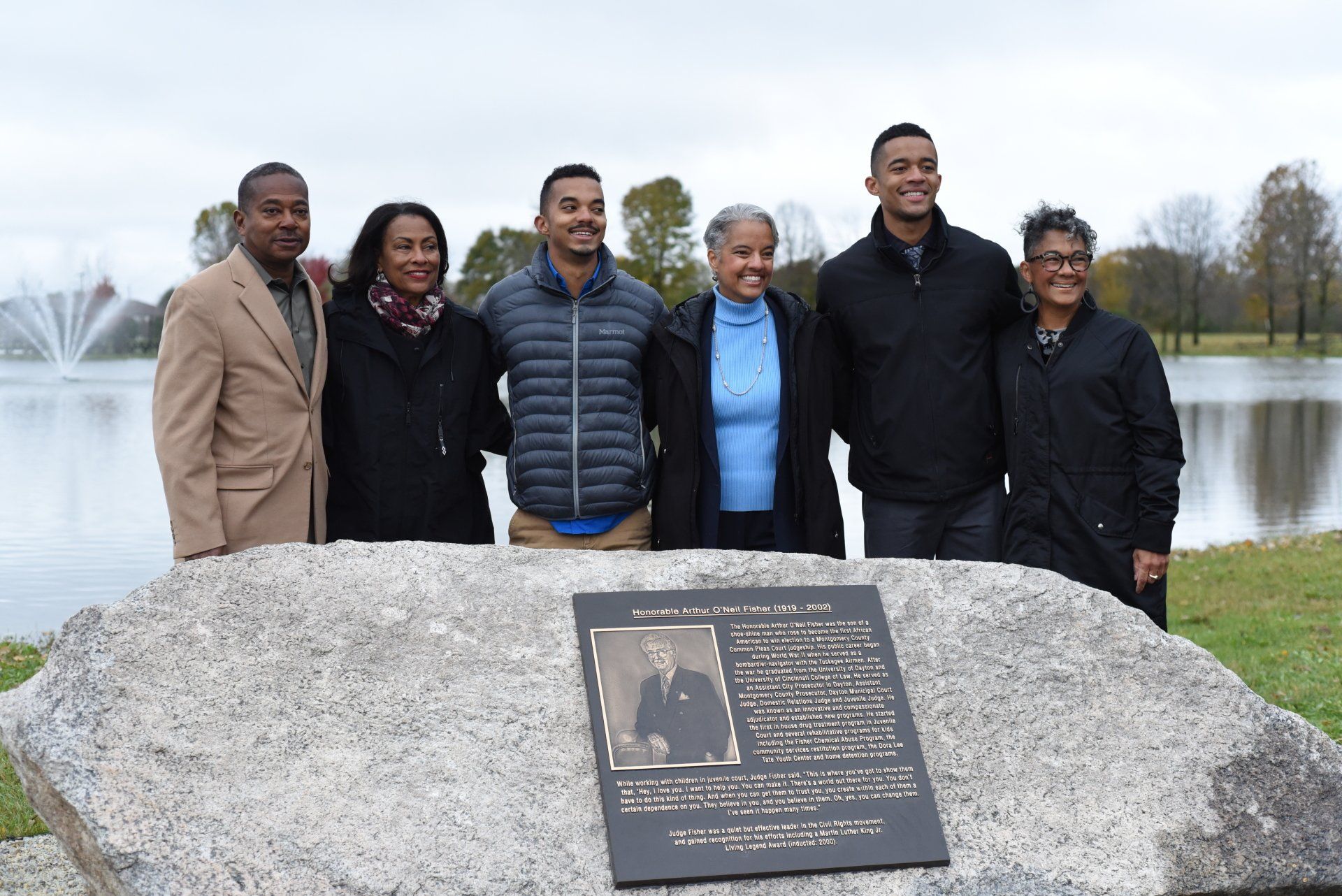 Group of people by a plaque, with a pond and fountain in the background.