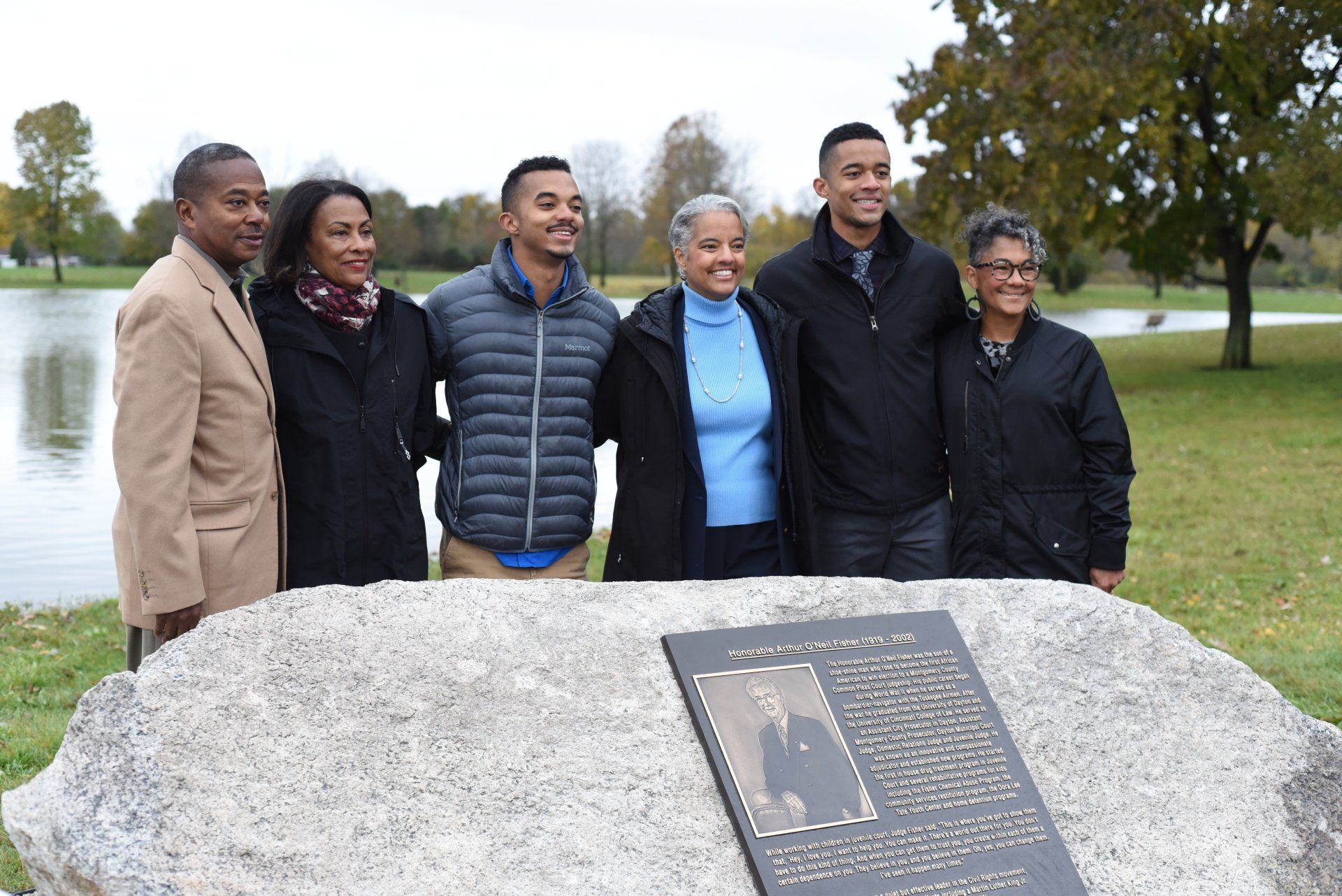 Family poses by plaque at a park with a lake. Several are smiling.