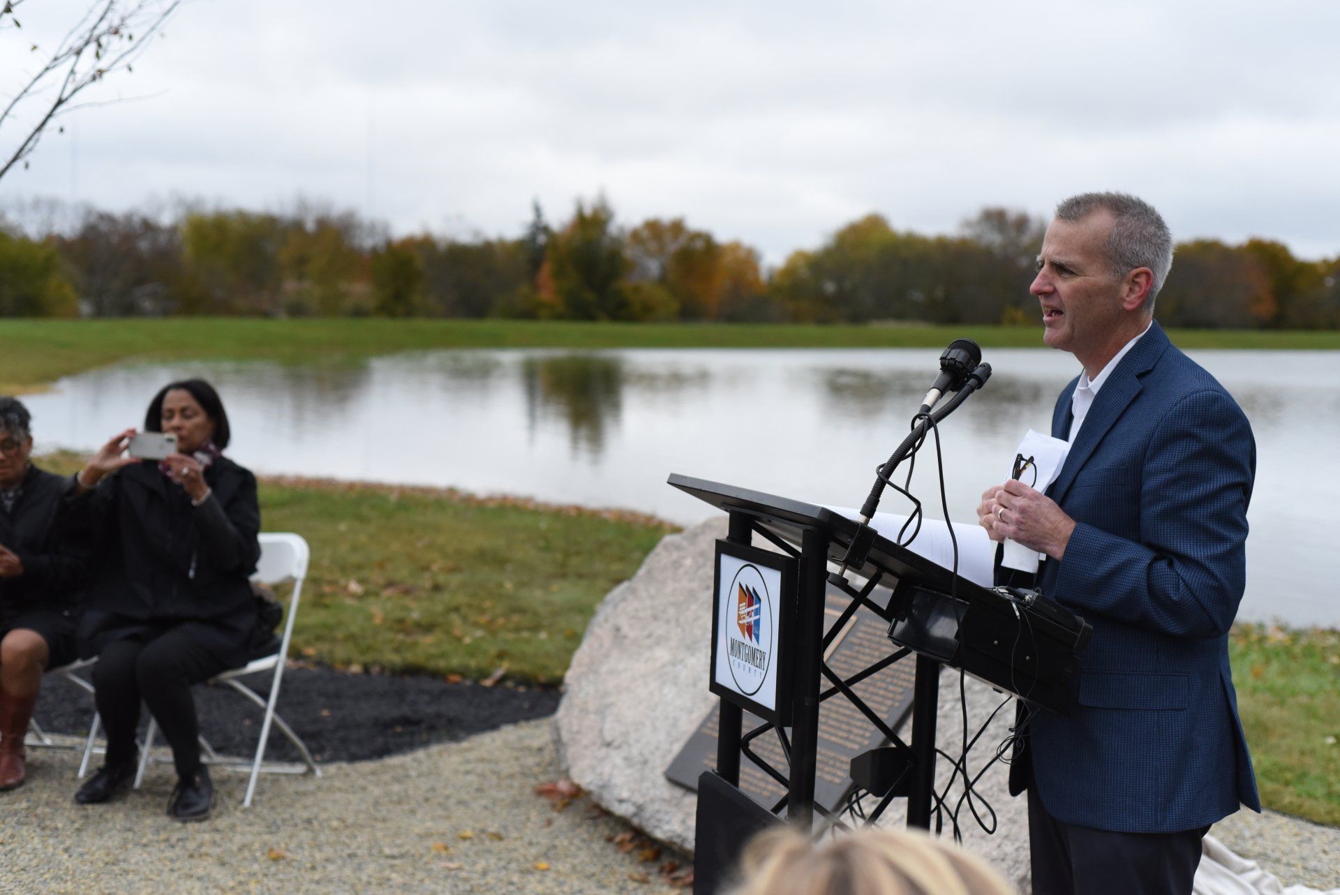 Man in suit speaks at podium near lake; woman takes a photo. Outdoors, cloudy.