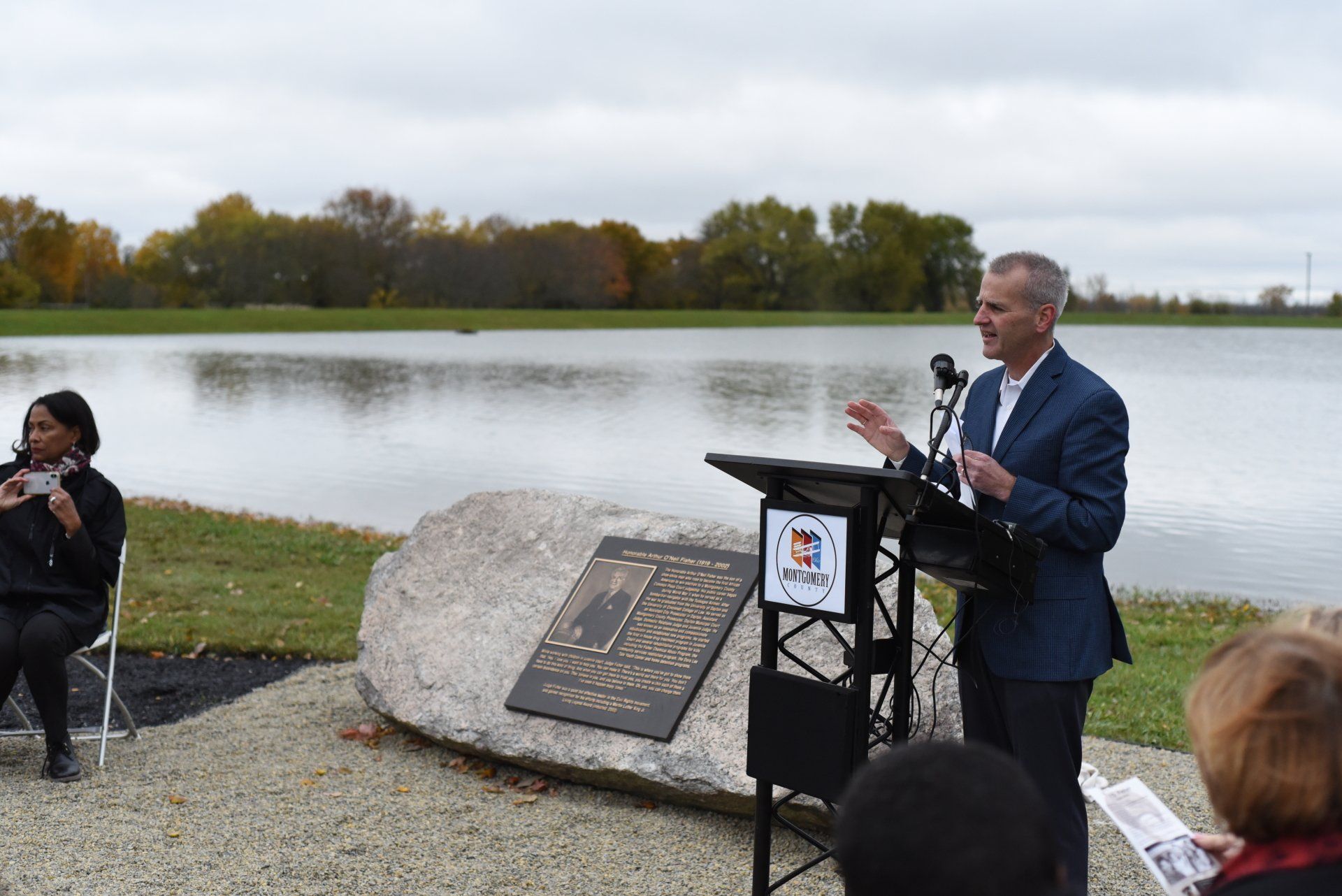 Man speaks at a podium by a lake; woman sits to the left. Memorial plaque on a large rock. Cloudy day.