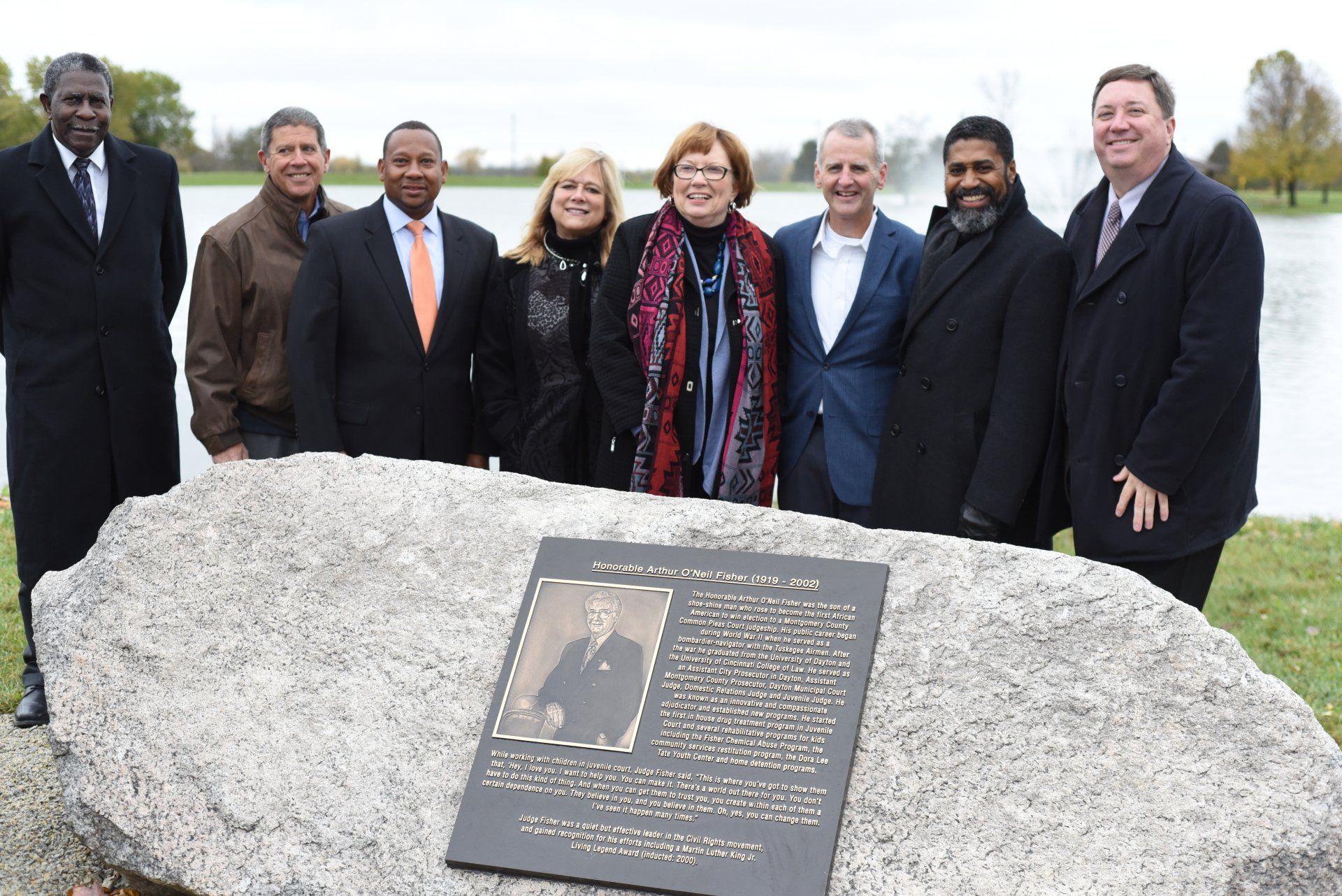 Group stands behind a memorial plaque near a lake; some are smiling and looking at the camera.