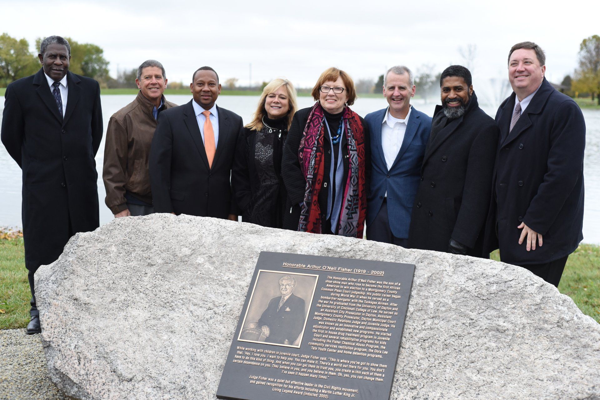 Group of people near a memorial plaque by a lake, standing outdoors.