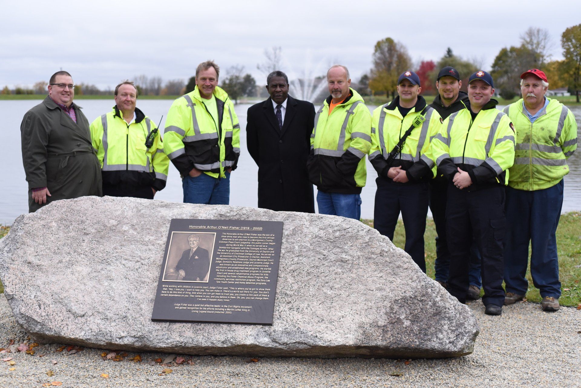 Group poses near a large rock with plaque at a park with a lake; autumn colors, cloudy skies.