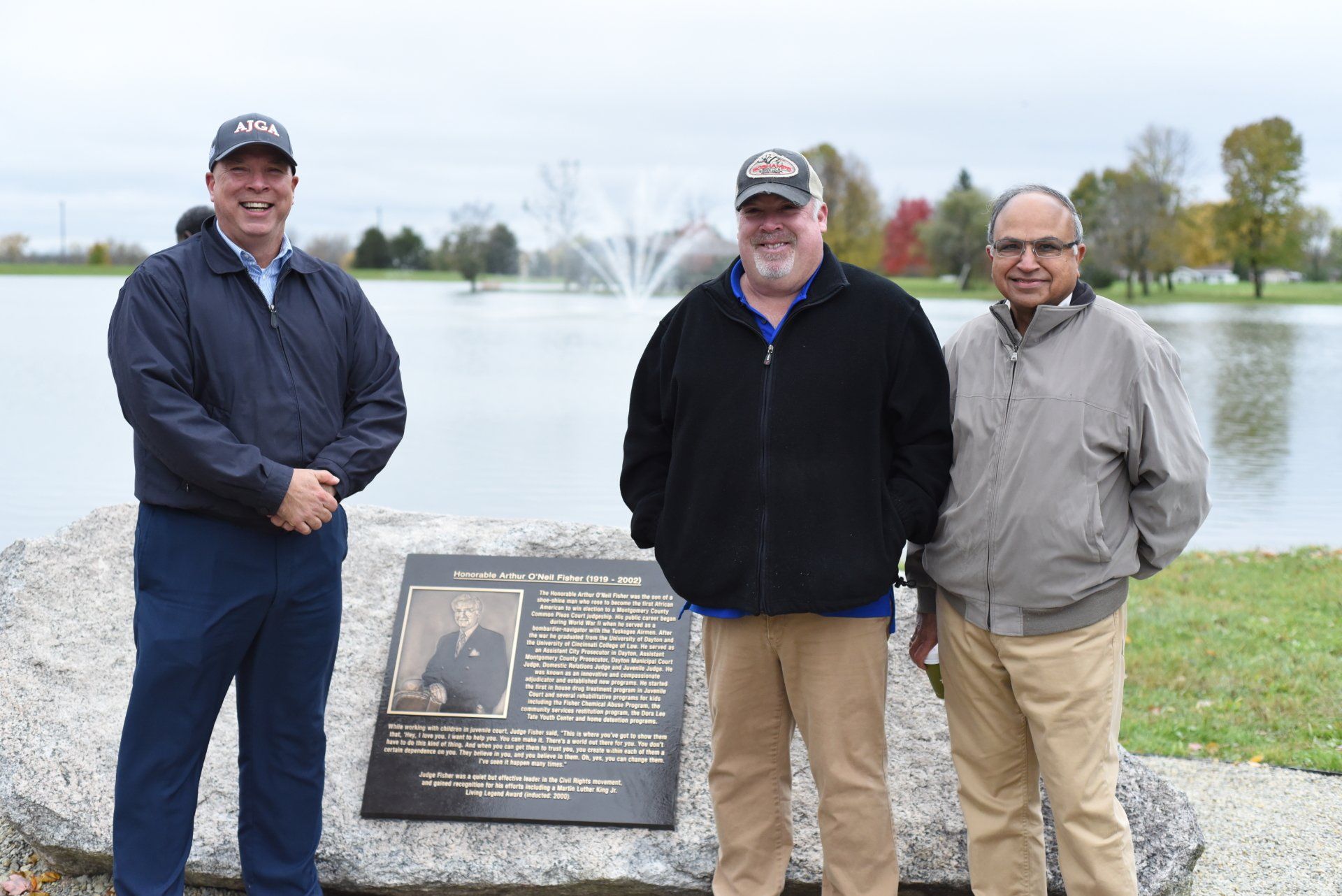 Three men stand by a dedication plaque next to a lake with a fountain.