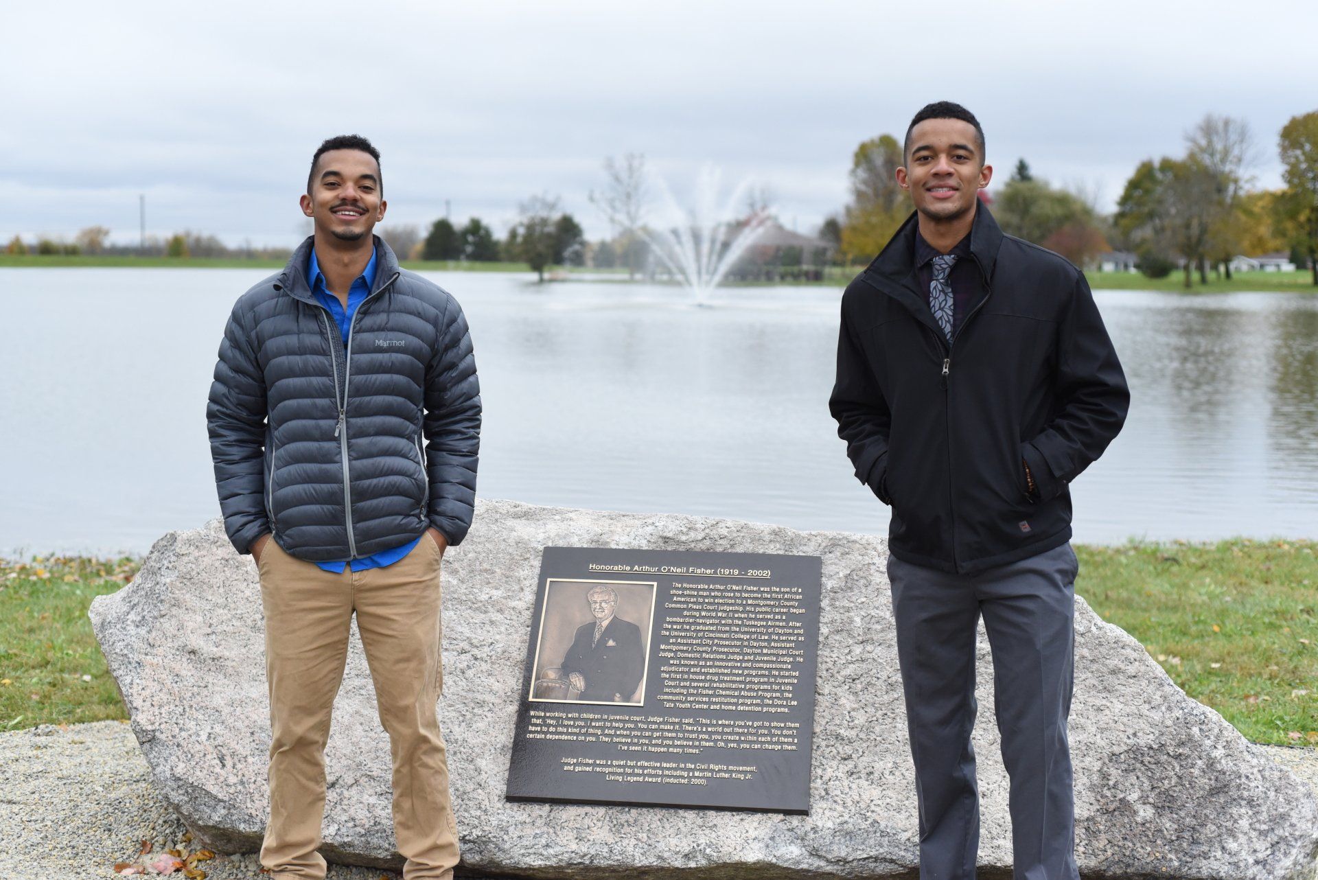 Two men standing by a monument, lake and fountain in the background. One wears a jacket, other a puffer coat.
