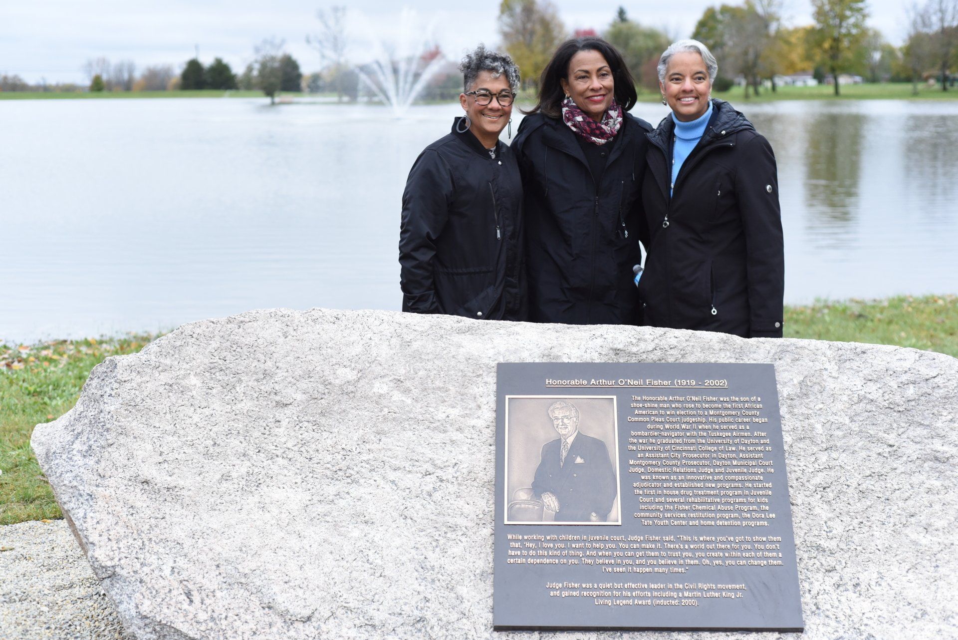 Three women stand by a plaque, lake in background. The plaque features a photo of a man and text.