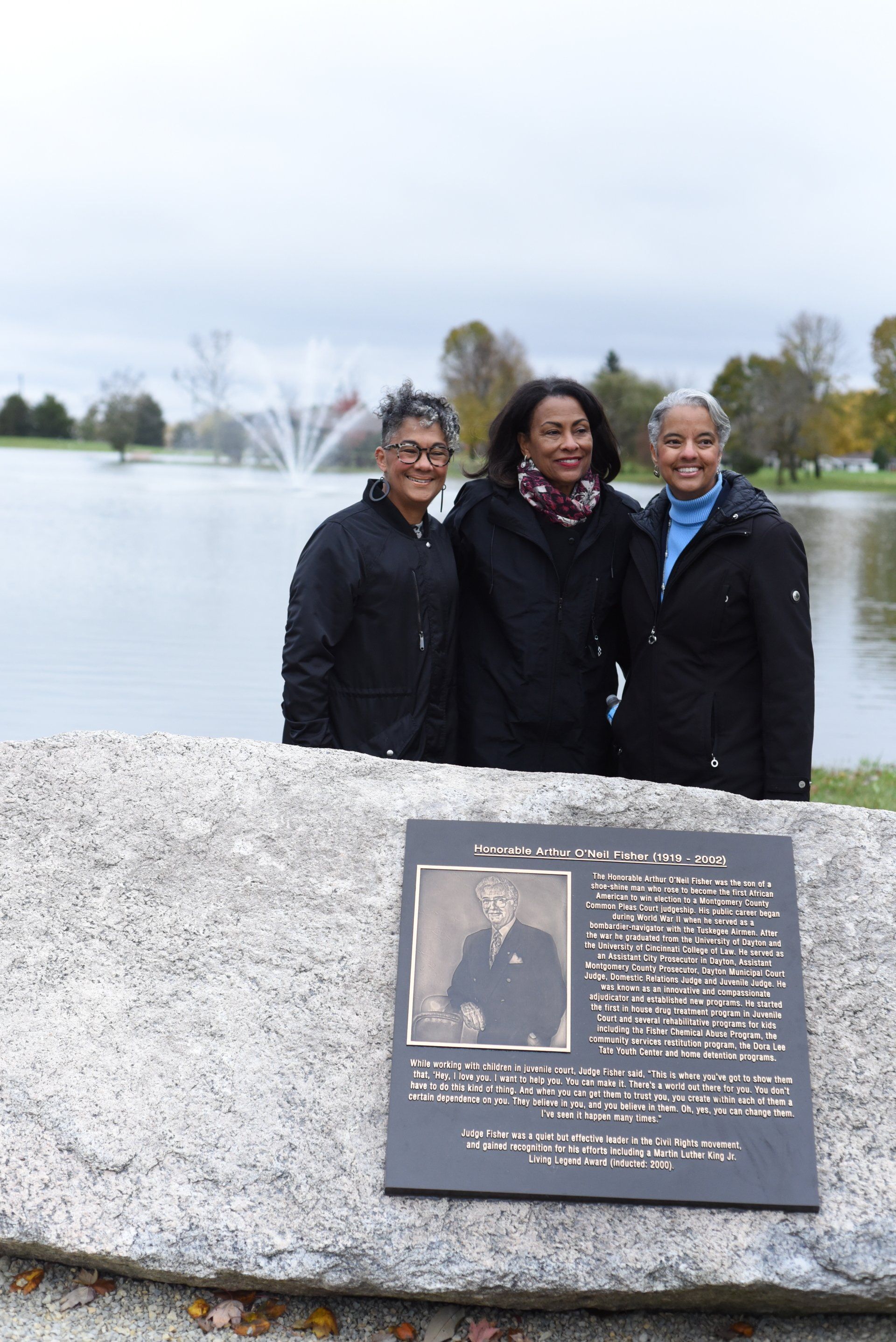 Three people stand near a memorial plaque by a lake, park setting; cloudy sky.