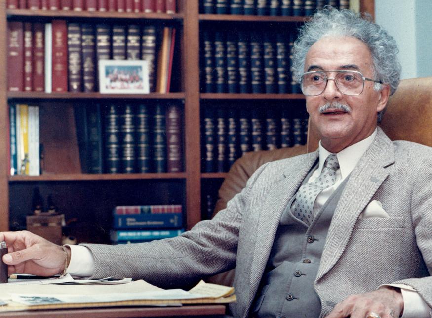 Man in a suit, eyeglasses, and curly hair sits at a desk in a library; bookshelves behind him.