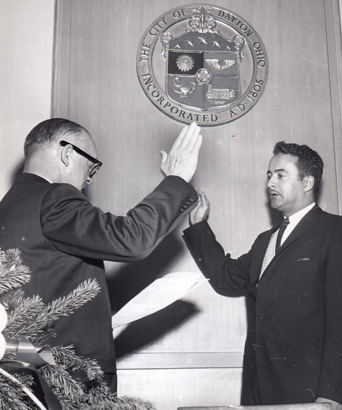 Two men, one in glasses, give high five in front of a city seal.