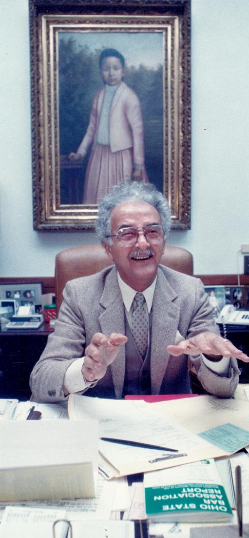 Man in a suit gestures while seated at a desk. A framed portrait hangs behind him.