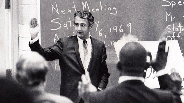 Man in suit with hand raised, addressing a group, likely a meeting. Blackboard in background, 1969.