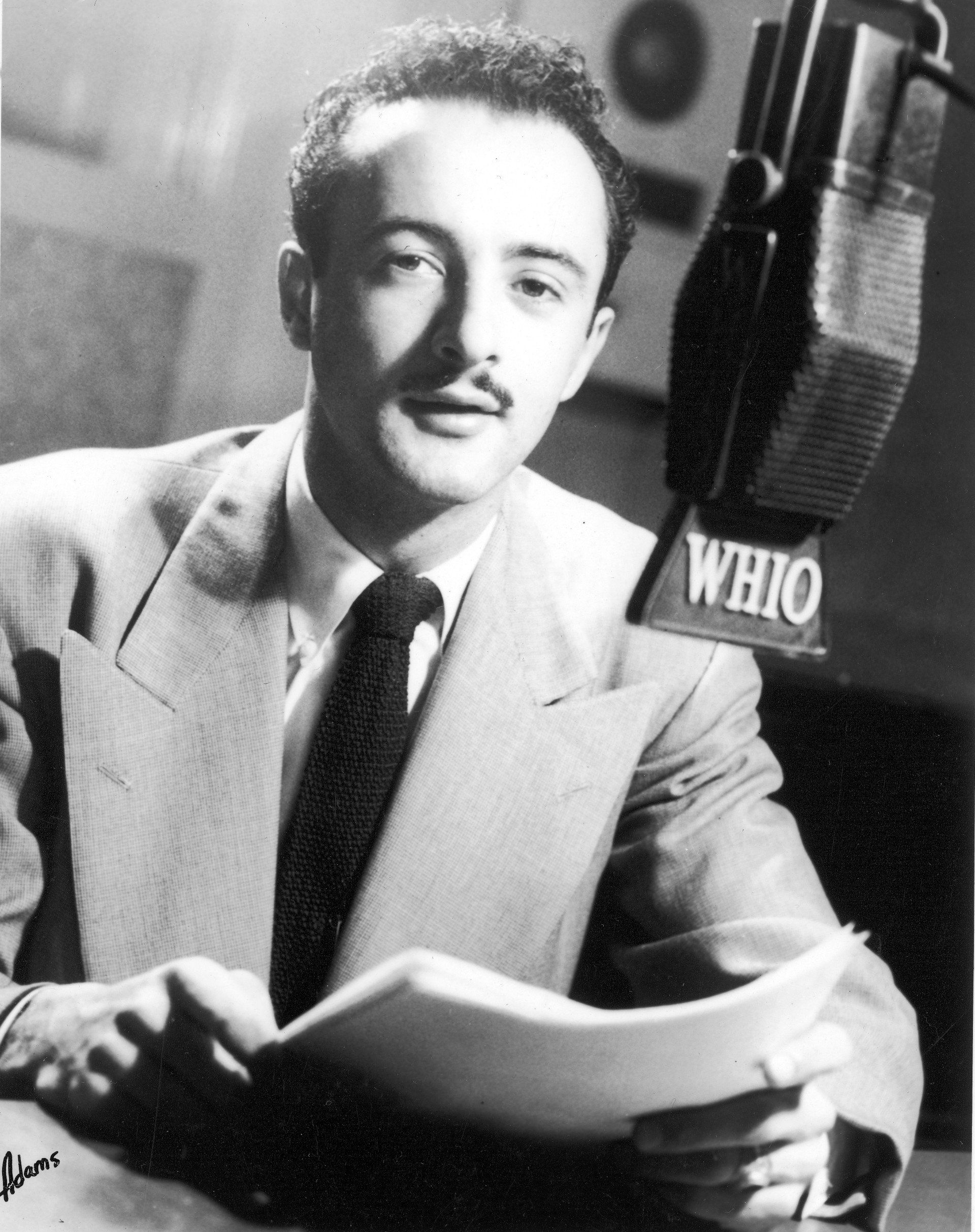 Man in a suit sits before a microphone, reading papers with the radio station's logo.