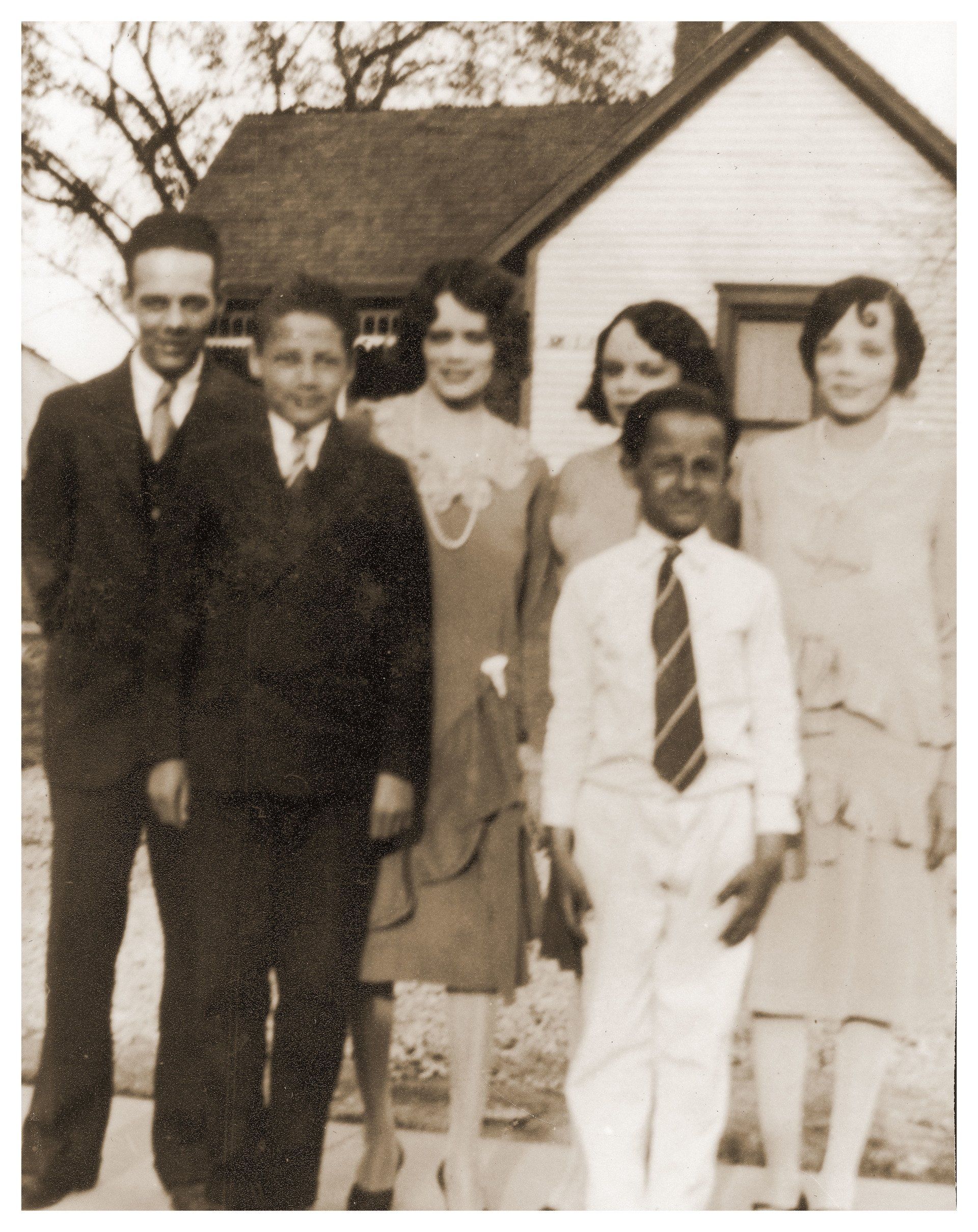 Group of six people posing in front of a house. Men, women, and children in formal clothing.