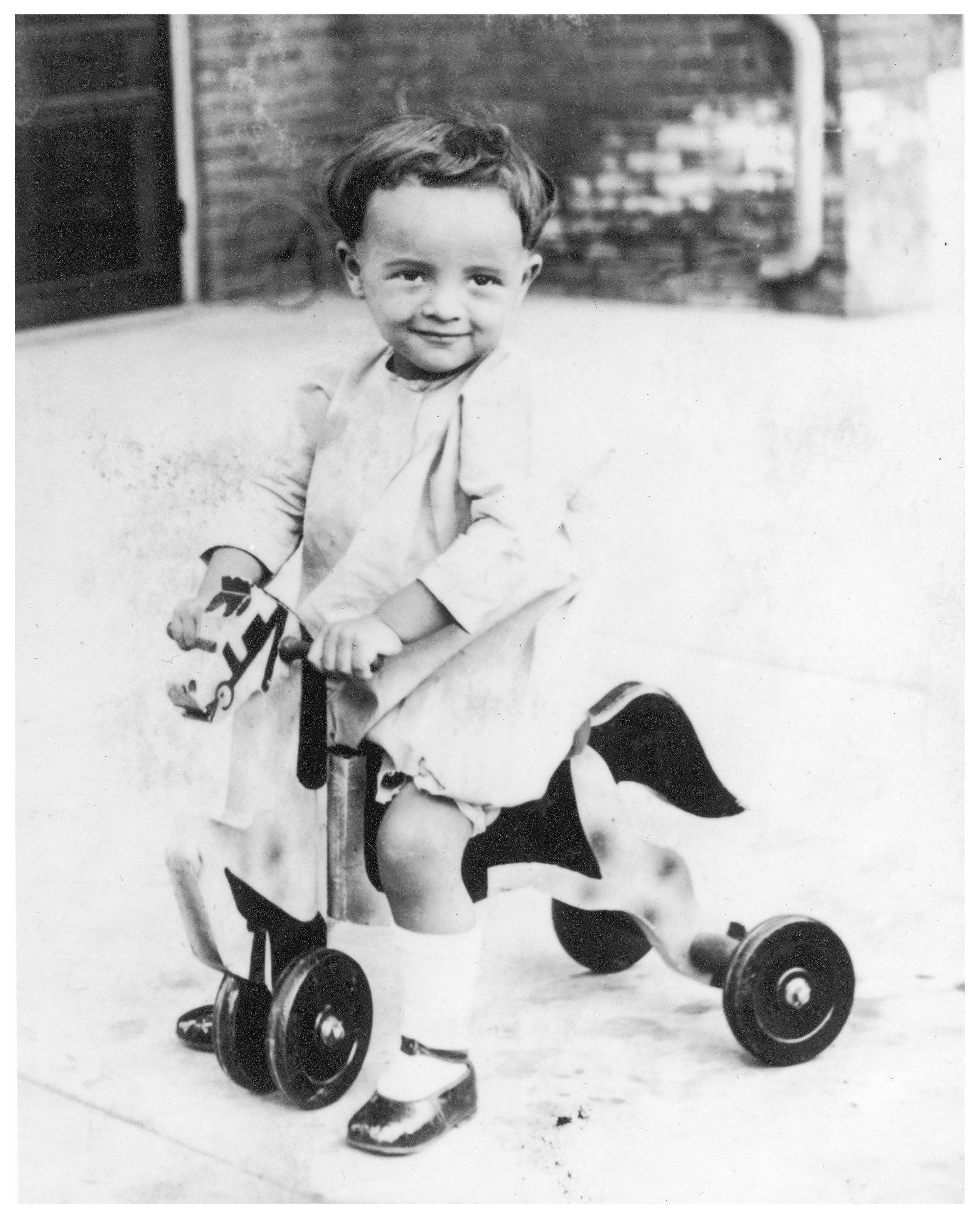 Smiling toddler on a tricycle shaped like a cow, wearing a light-colored dress and socks, outside.