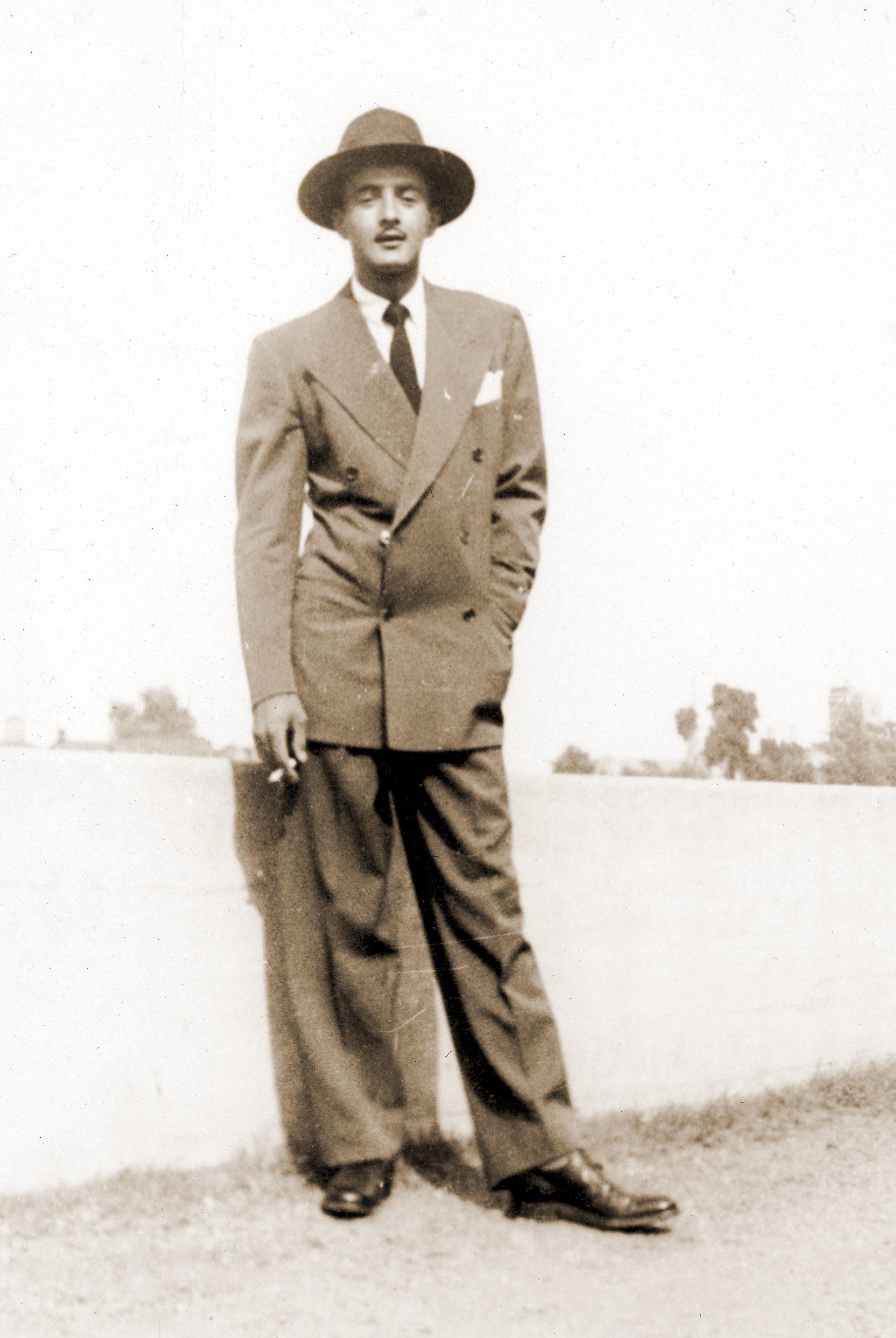 Man in a double-breasted suit and hat, smoking, leaning against a wall outdoors.