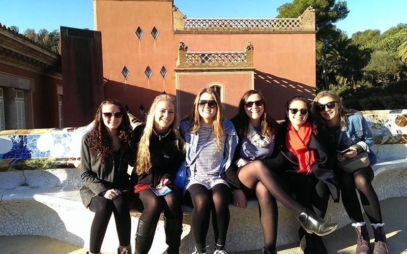 Six young women with sunglasses, sitting on a colorful bench in front of a red building.
