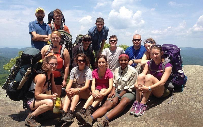 Group of hikers atop a mountain, smiling. Sunny day, backpacks visible.