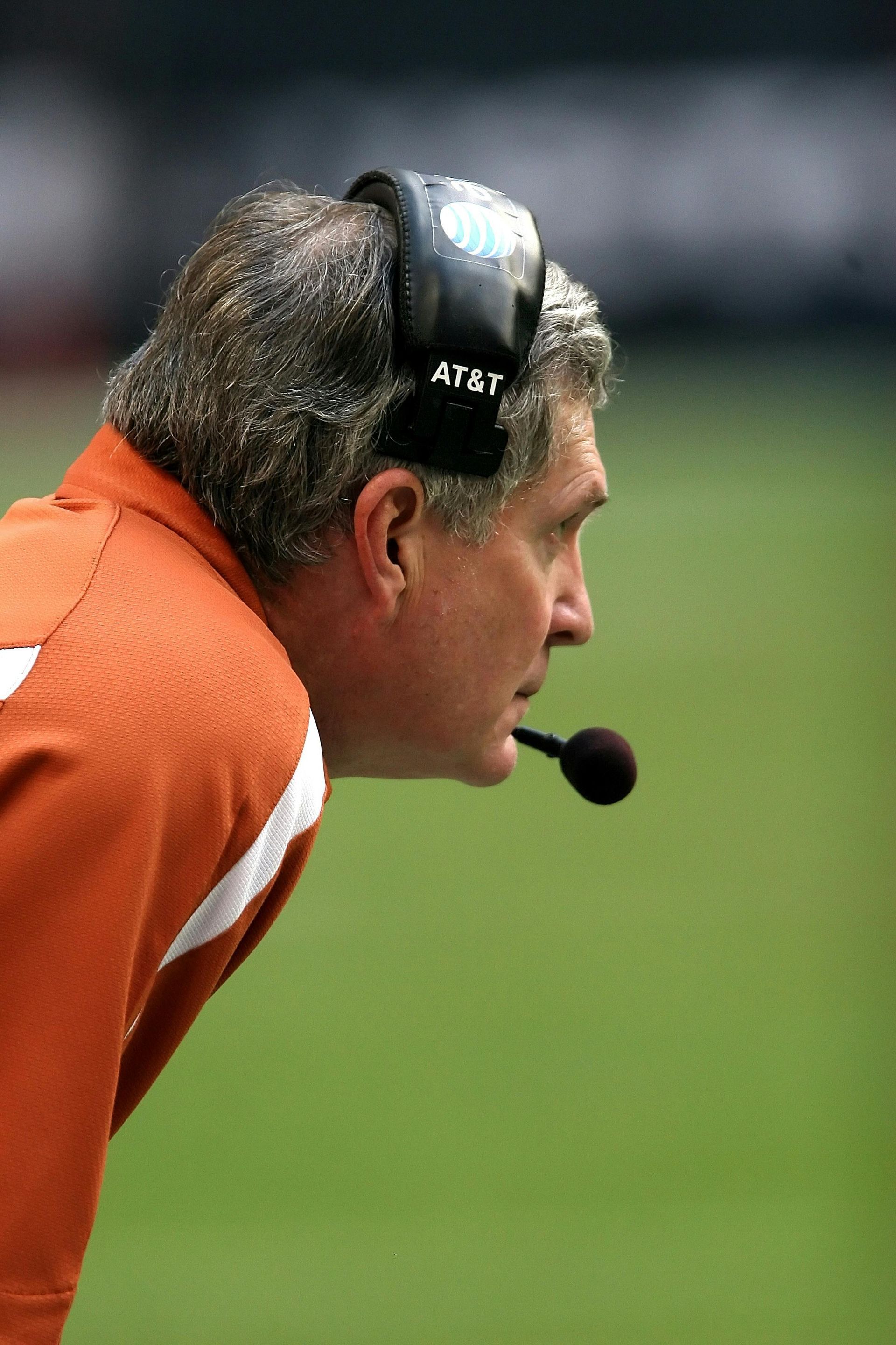 Coach in burnt orange shirt, headset with microphone, watching a football game on a green field.