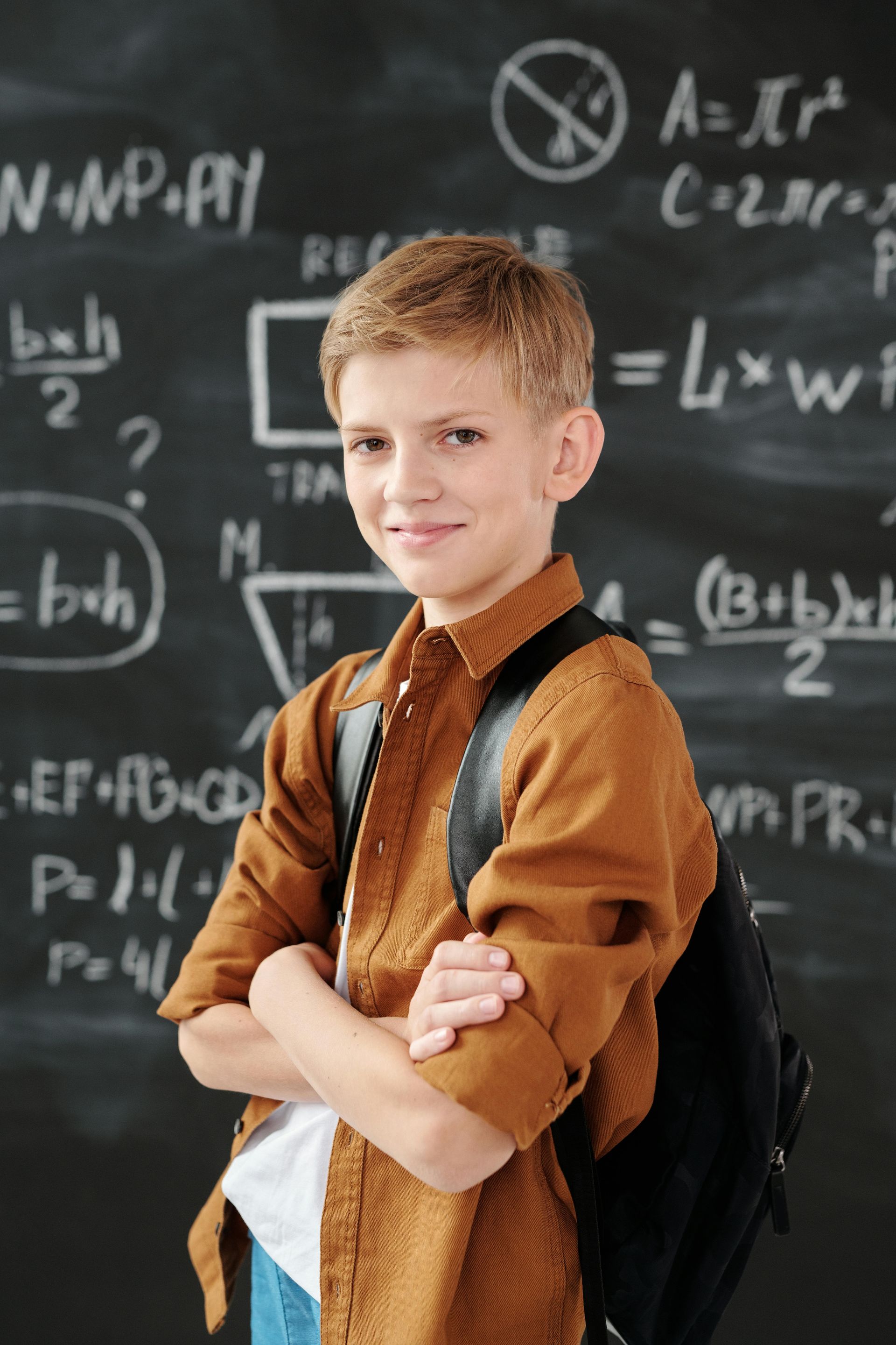 Boy with arms crossed, backpack, smiling in front of a chalkboard filled with math formulas.