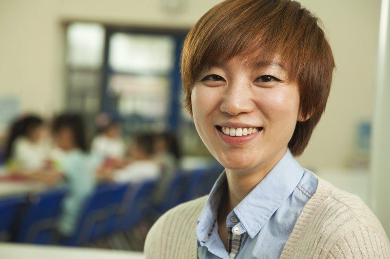 Smiling Asian woman in a classroom, wearing a cardigan and light blue shirt, with children blurred in the background.