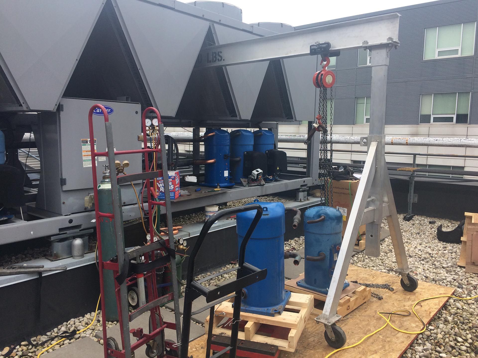 HVAC equipment on a rooftop. Blue cylinders, a metal cart with tools, a hoist, and dark triangular structures.