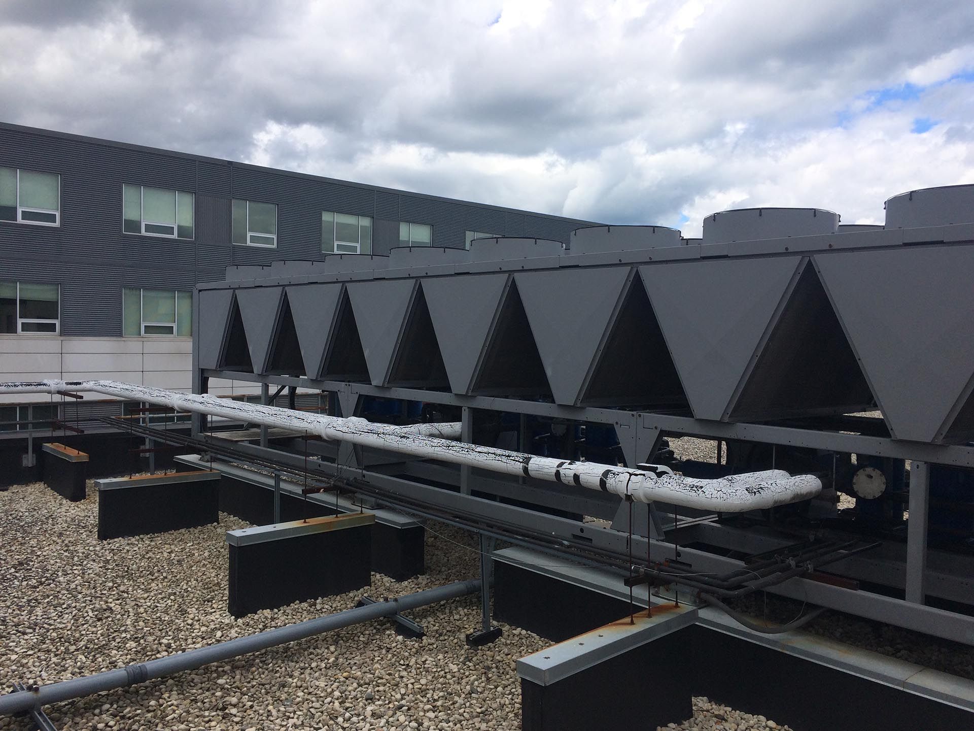 Roof-mounted industrial cooling units with triangular vents and connected pipes, against a gray building and cloudy sky.