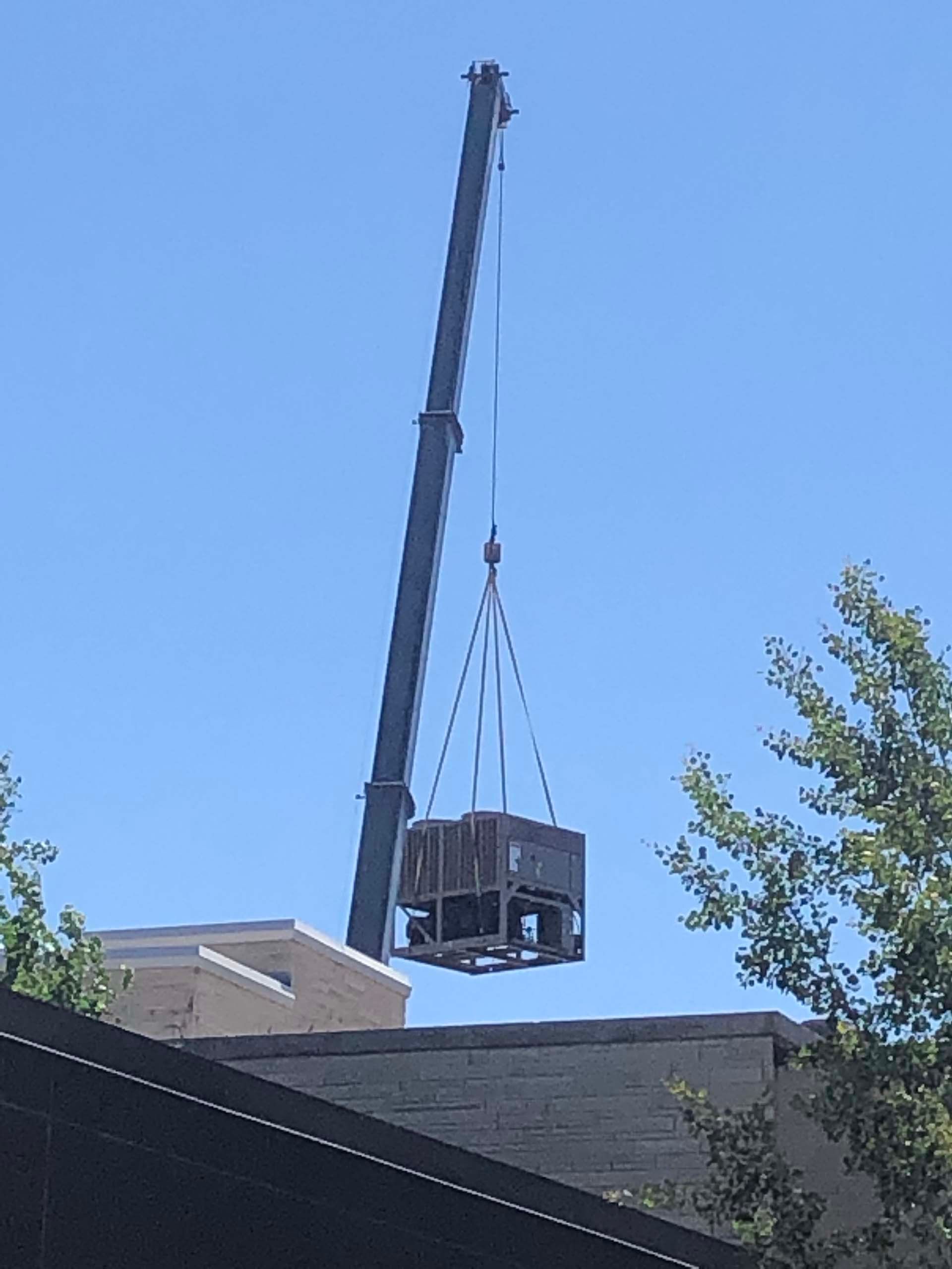 A crane lifts a metal crate over a building's roof against a clear blue sky.