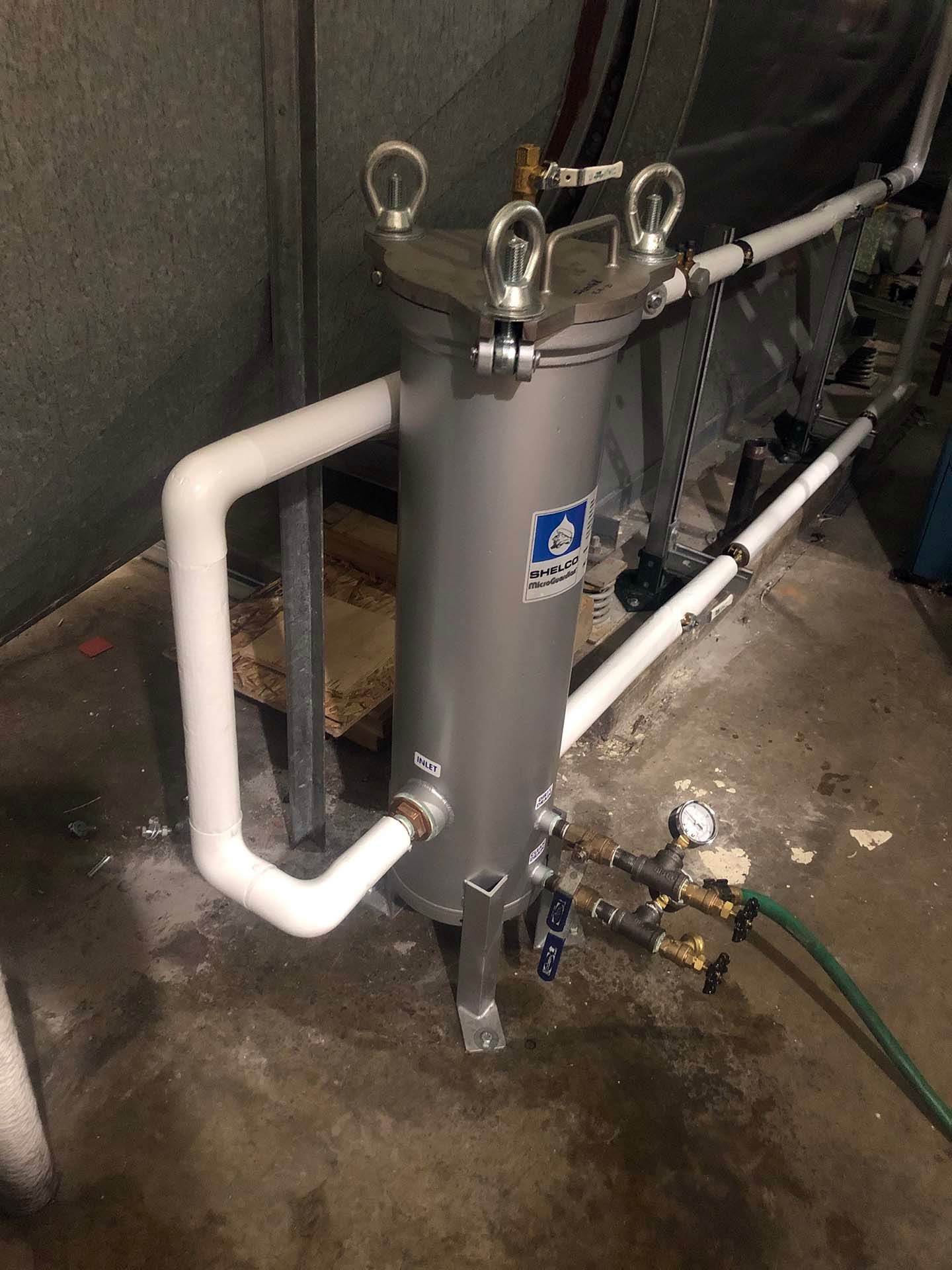 A silver water filter with white PVC pipes, metal fittings, and a connected green hose in a basement setting.