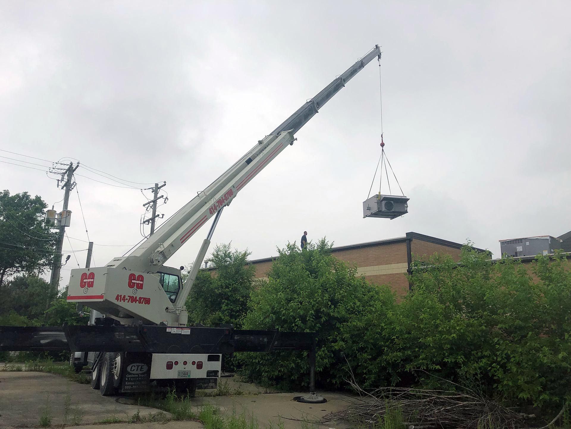 Crane lifting an object near a building with overgrown bushes. Cloudy sky.