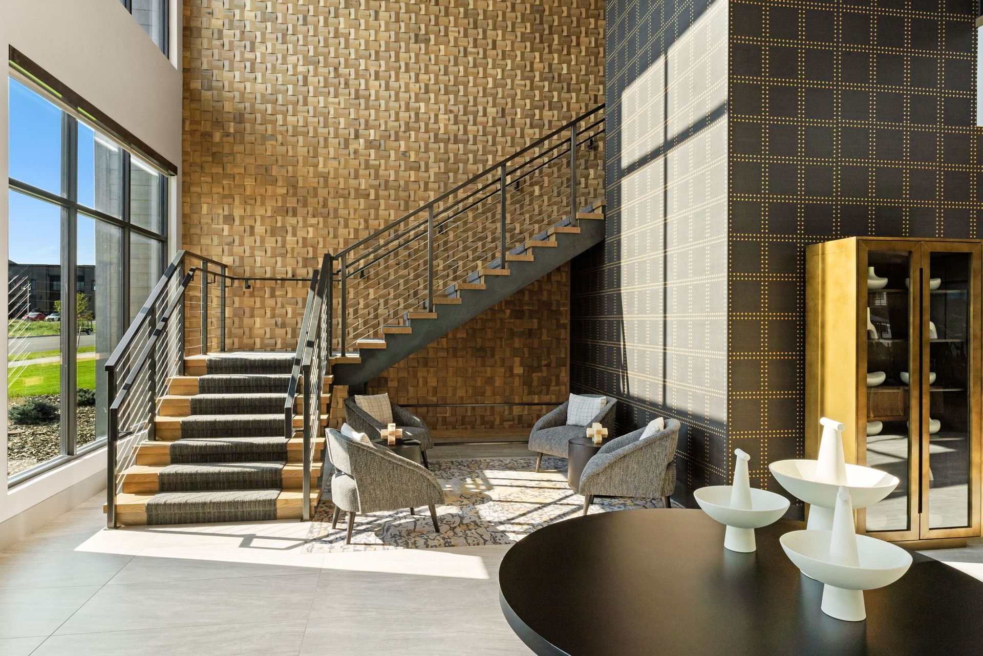 Lobby with staircase, wicker seating, and a large black table. Textured walls, tall windows, and a gold cabinet.