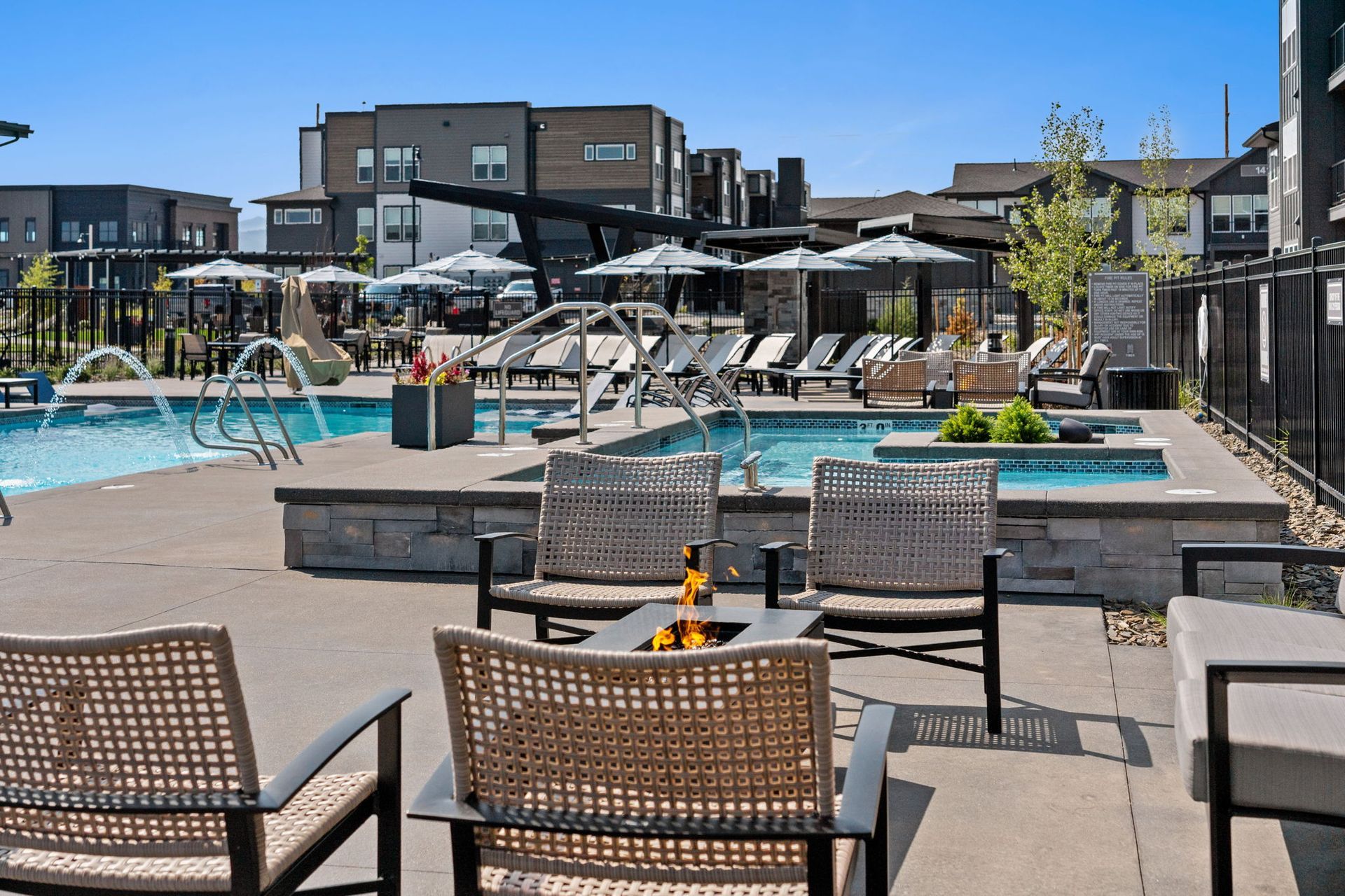 Pool area with lounge chairs, a fire pit, and a jacuzzi. Modern buildings in the background.
