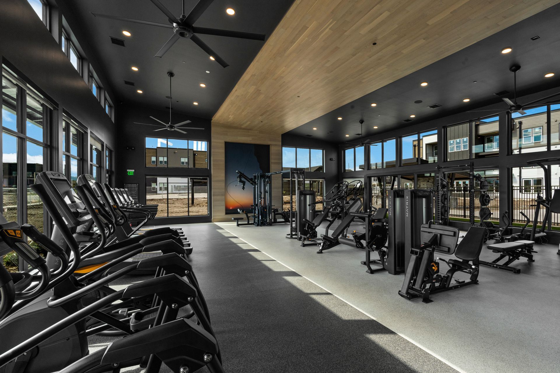 Gym with cardio machines and weight equipment, large windows, and dark ceiling.