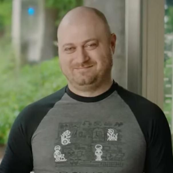 Solomon, a middle aged, medium build, bald, white man smiling in front of a window while wearing a grey and black shirt