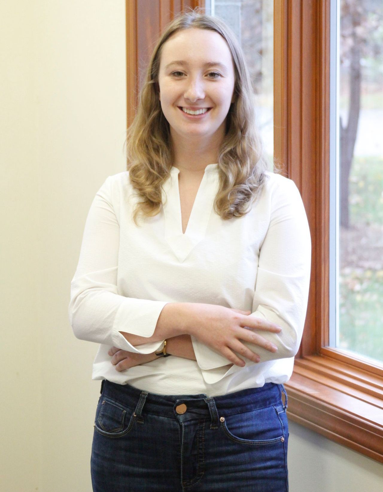 A woman in a white shirt and jeans is standing in front of a window with her arms crossed.