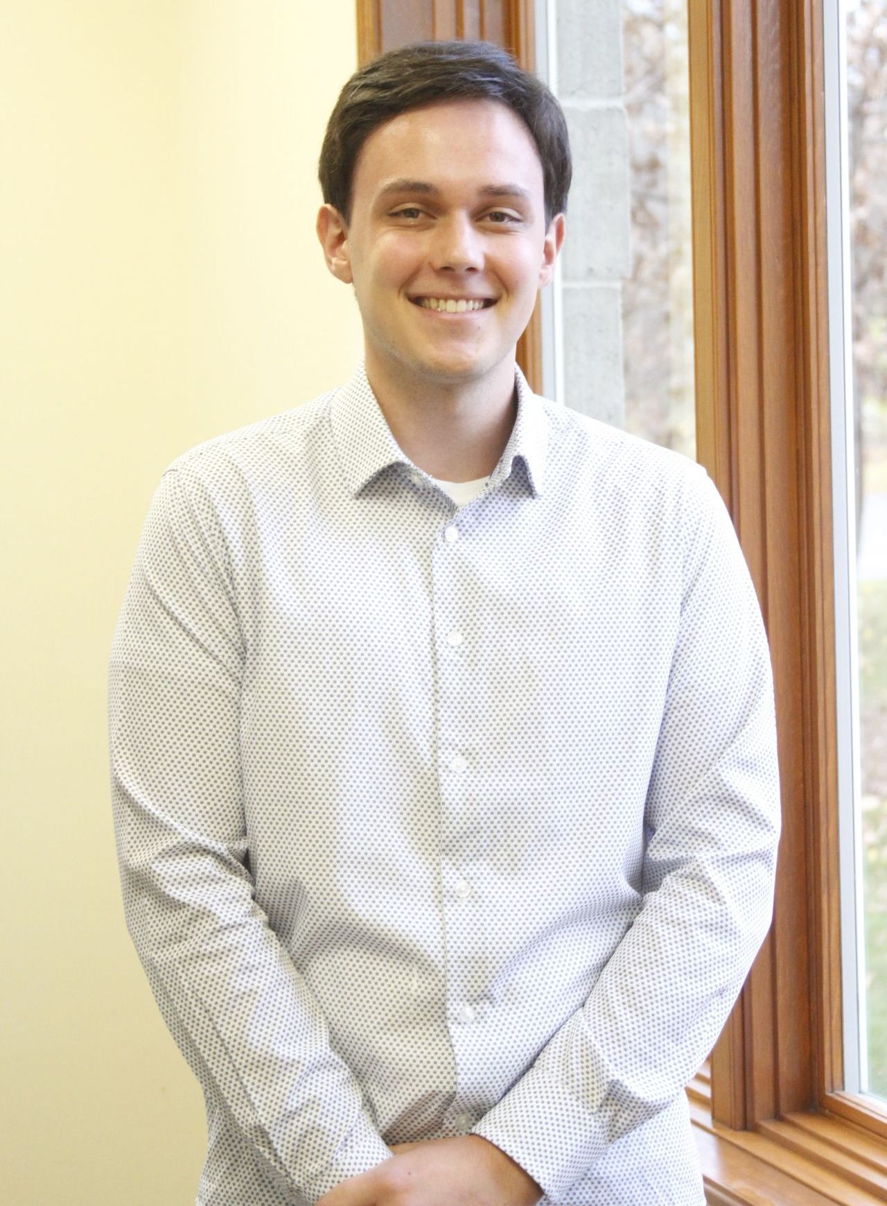 A young man in a white shirt is standing in front of a window.