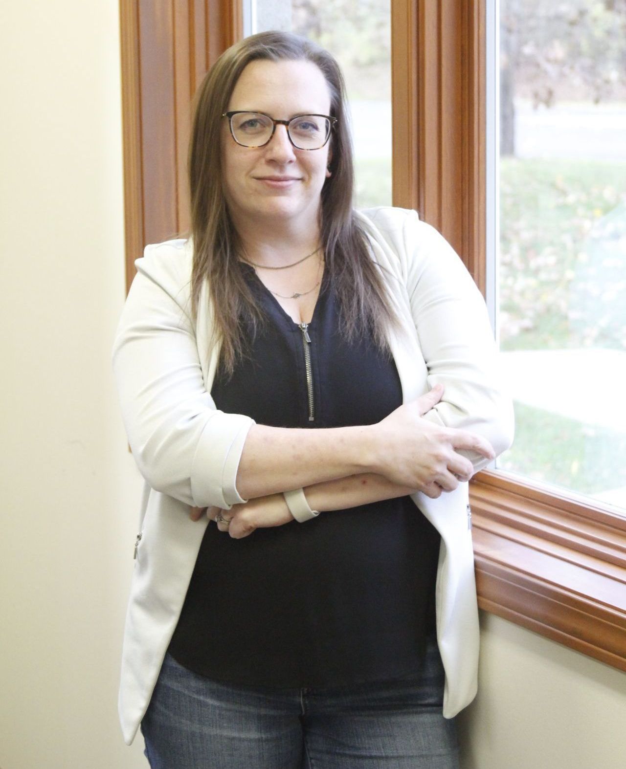 A woman wearing glasses is standing in front of a window with her arms crossed.