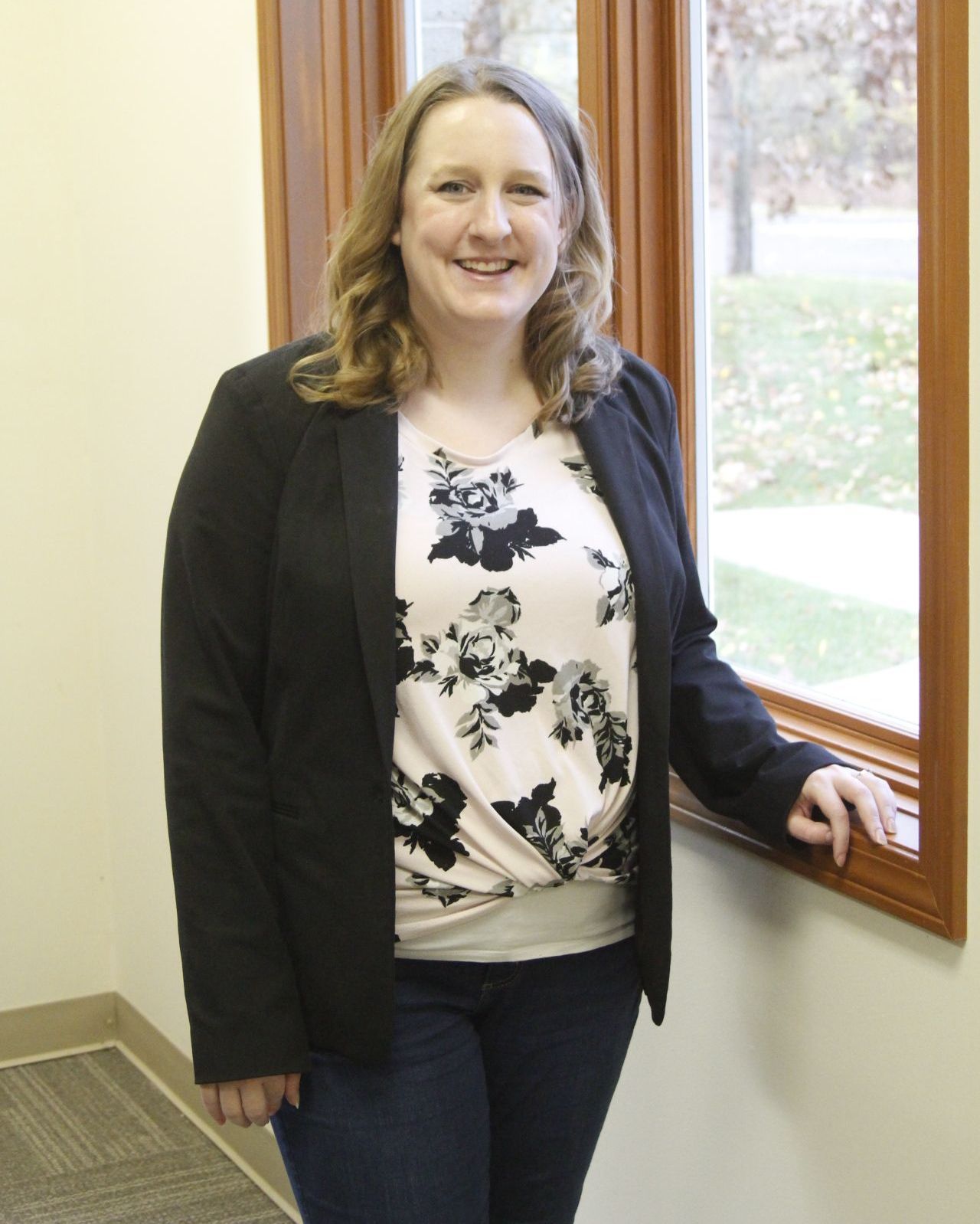 A woman in a floral shirt and a black jacket is standing next to a window.