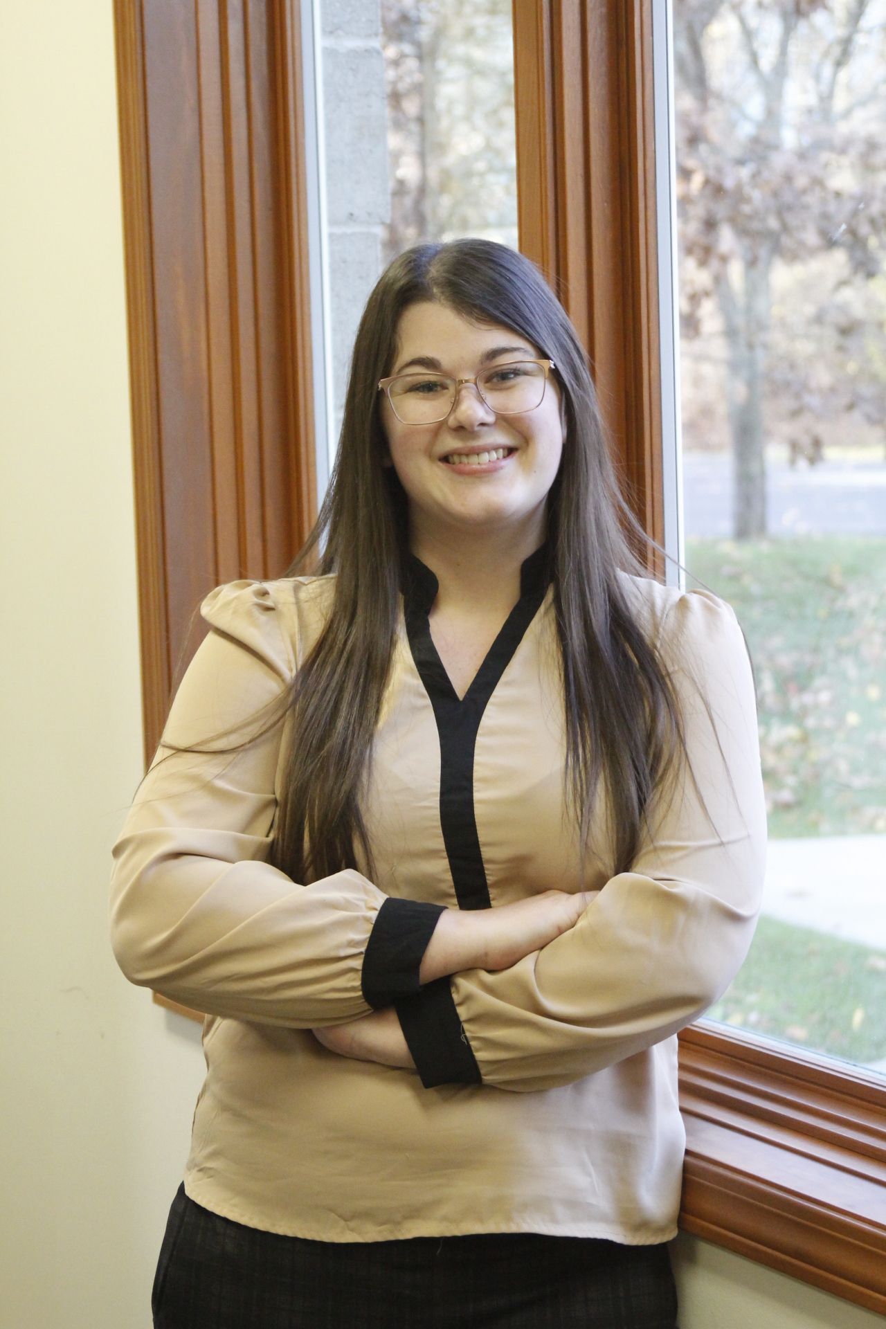 A woman is standing in front of a window with her arms crossed and smiling.