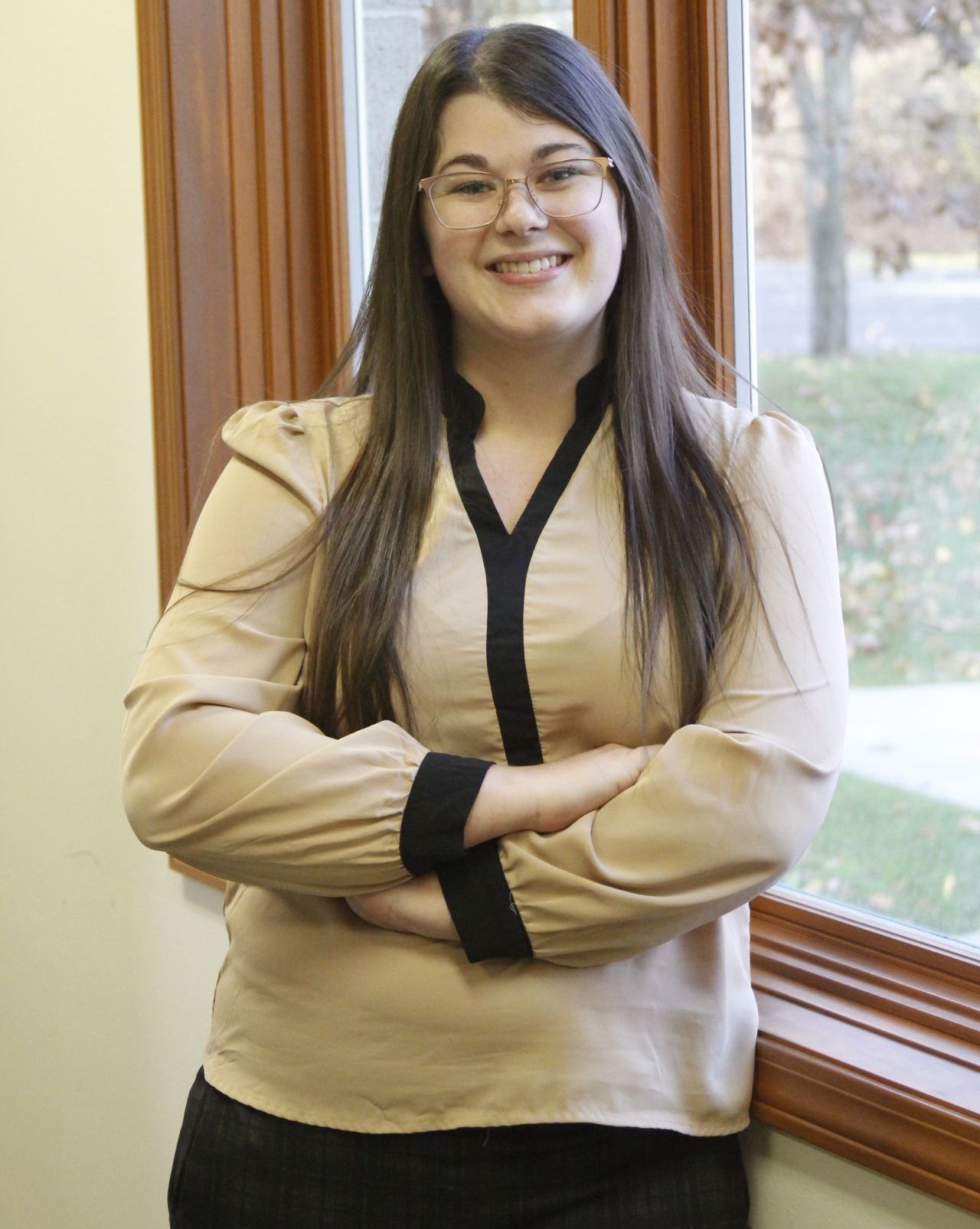 A woman is standing in front of a window with her arms crossed and smiling.