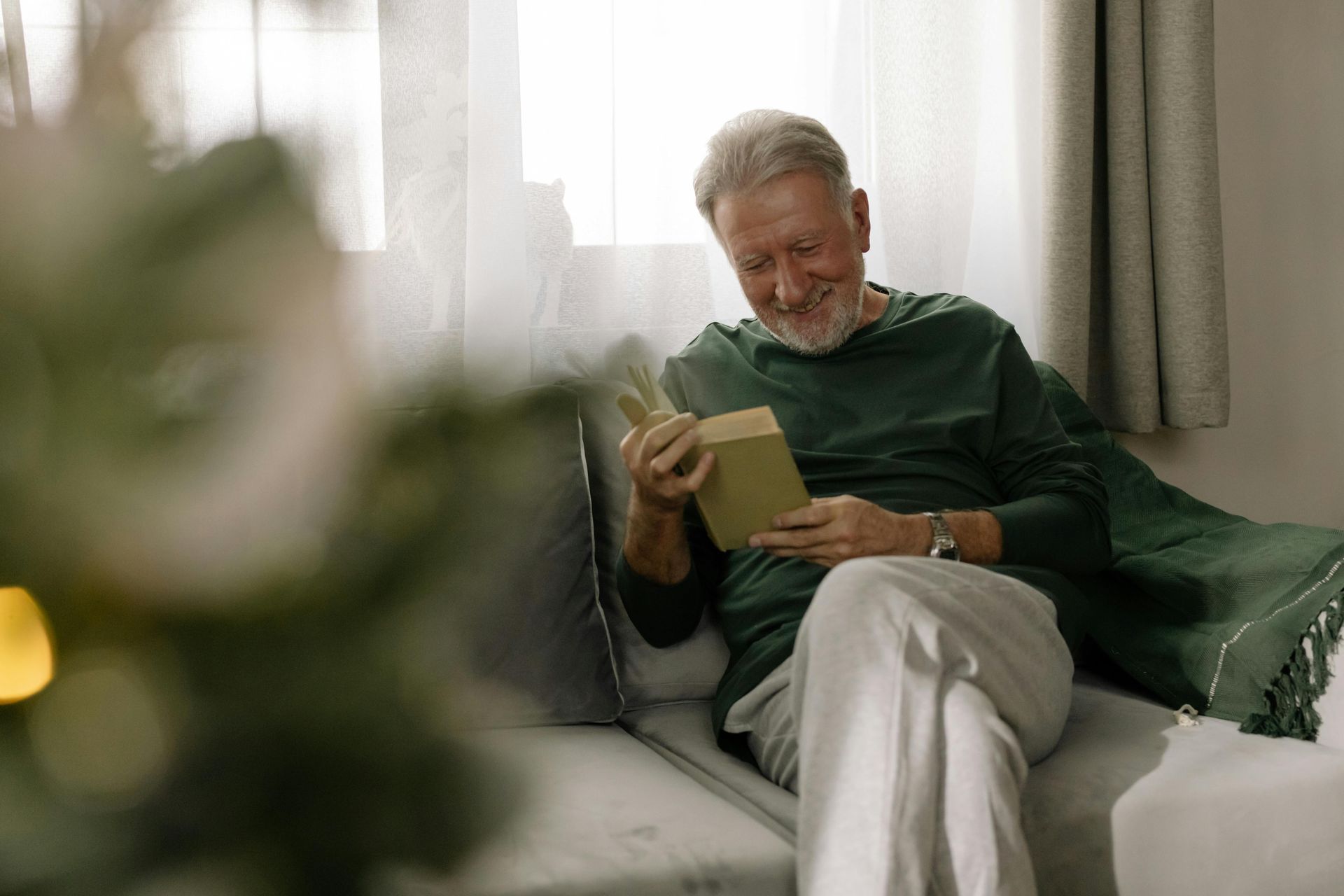 An elderly man is sitting on a couch reading a book.