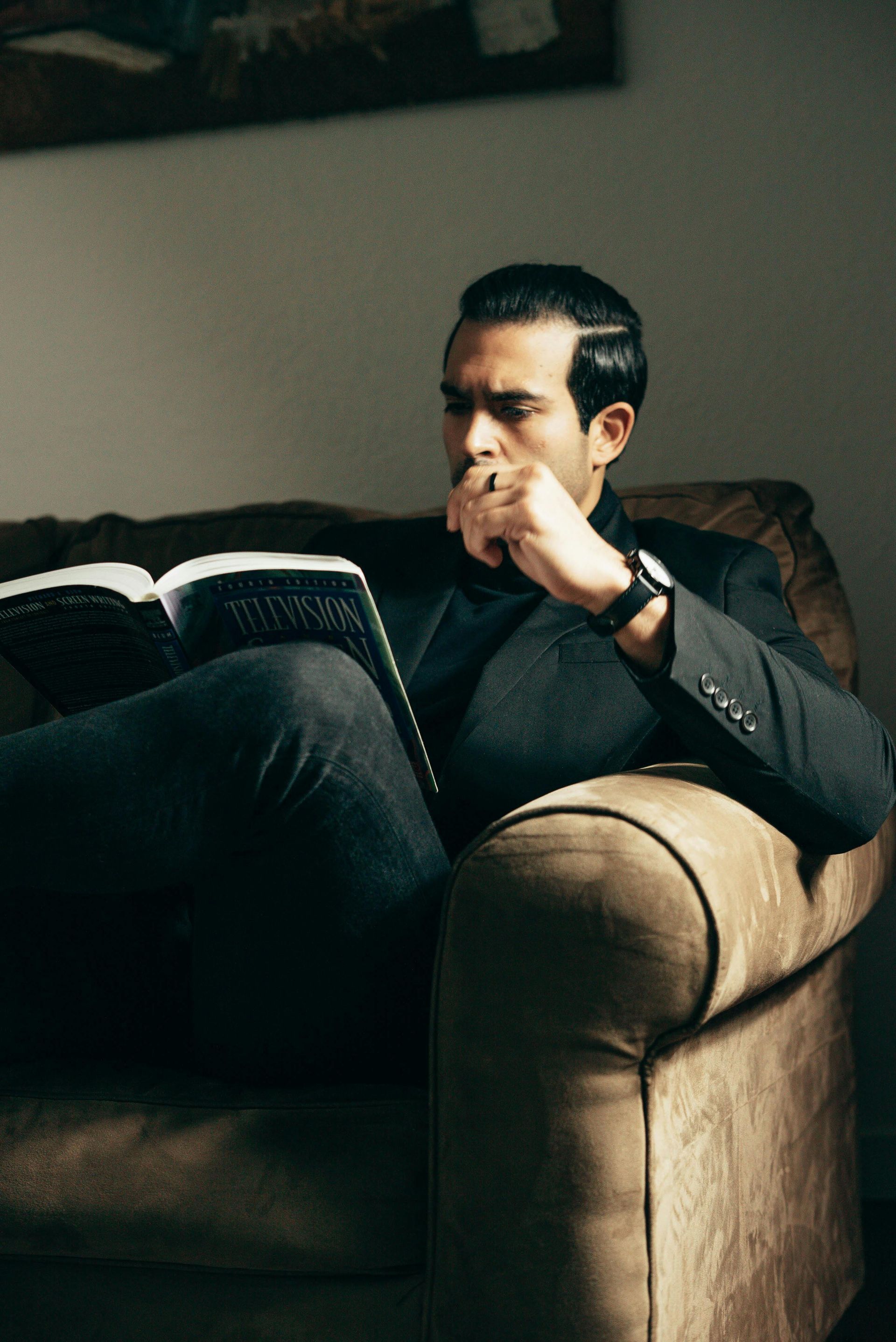 A man in a suit is sitting on a couch reading a book.