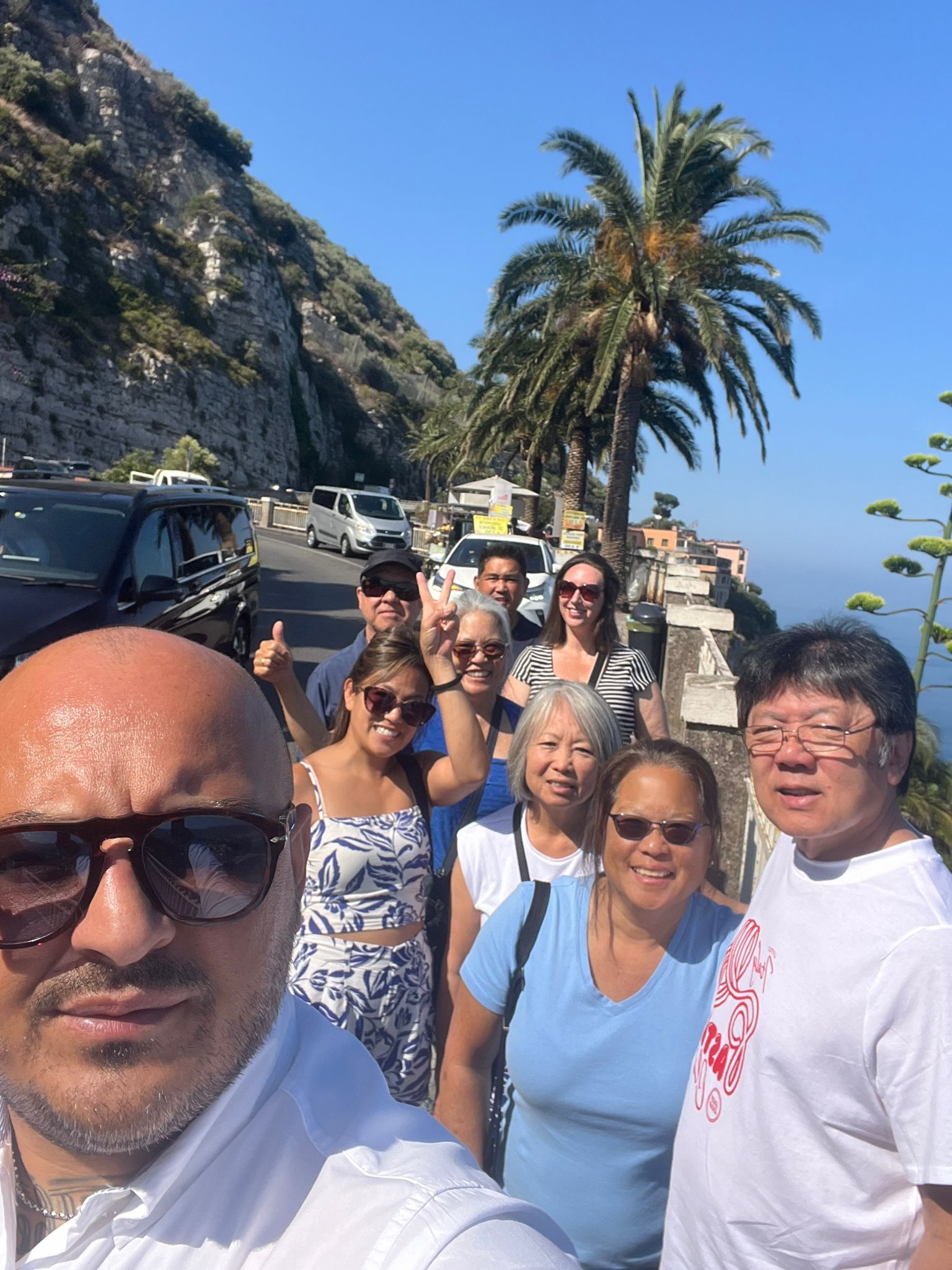 Selfie di gruppo su una strada costiera. Persone sorridenti vicino ad auto parcheggiate, palme e vista sull'oceano. Giornata di sole.