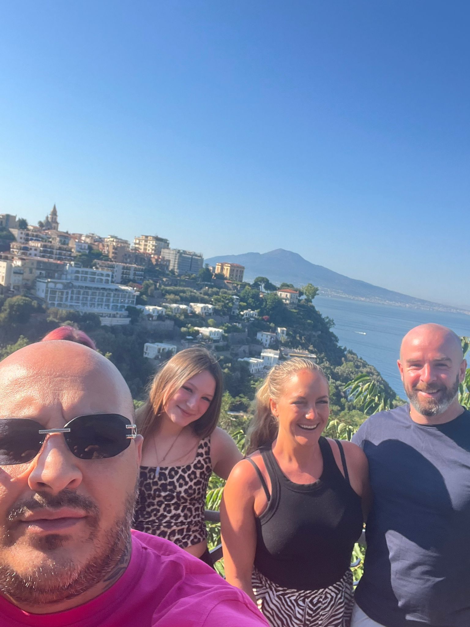 Quattro persone posano per una foto con una città costiera sullo sfondo. Giornata di sole splendente.