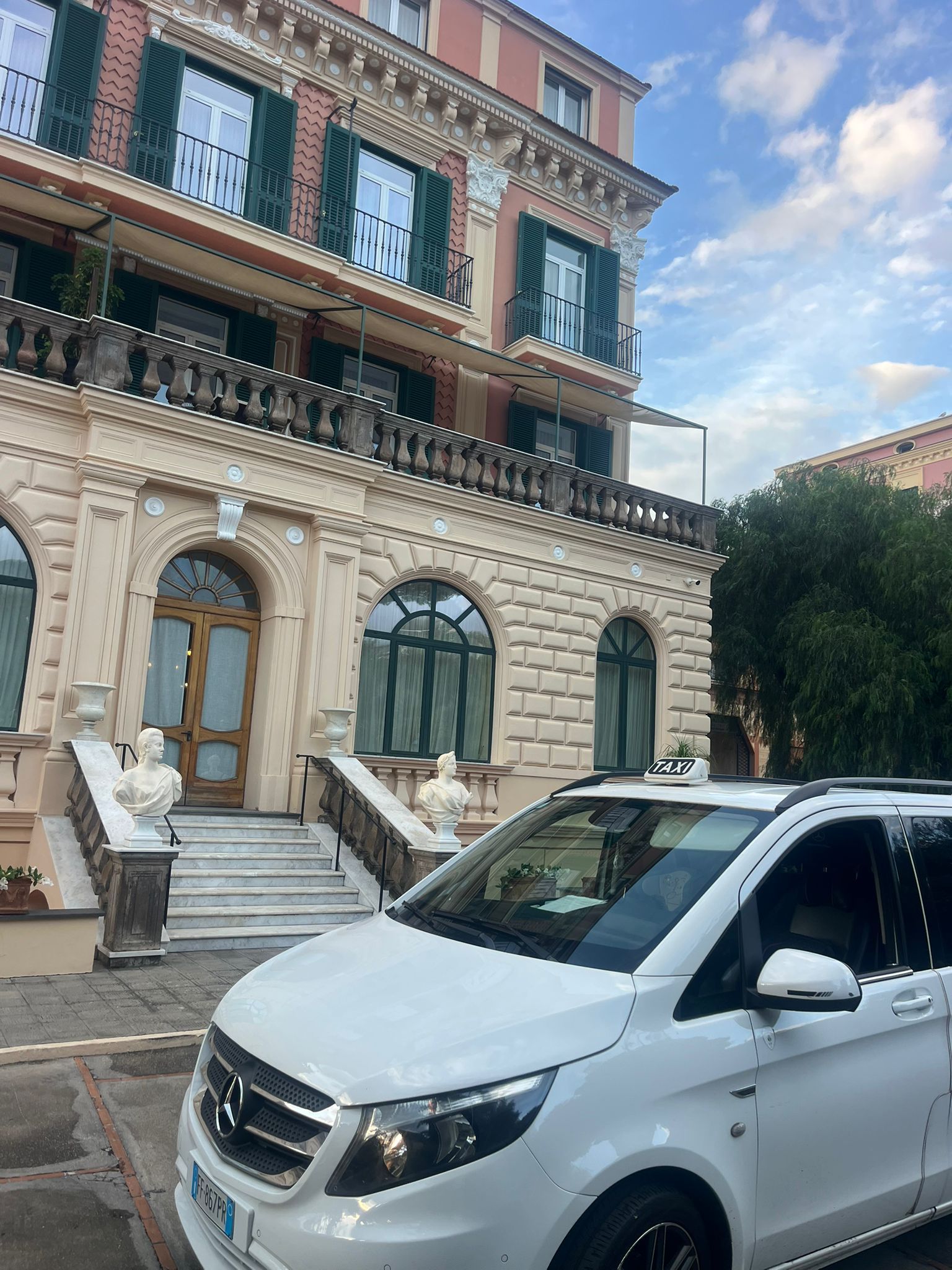 Furgone bianco parcheggiato fuori da un imponente edificio con facciata in pietra, balcone e dettagli decorati. Cielo azzurro.